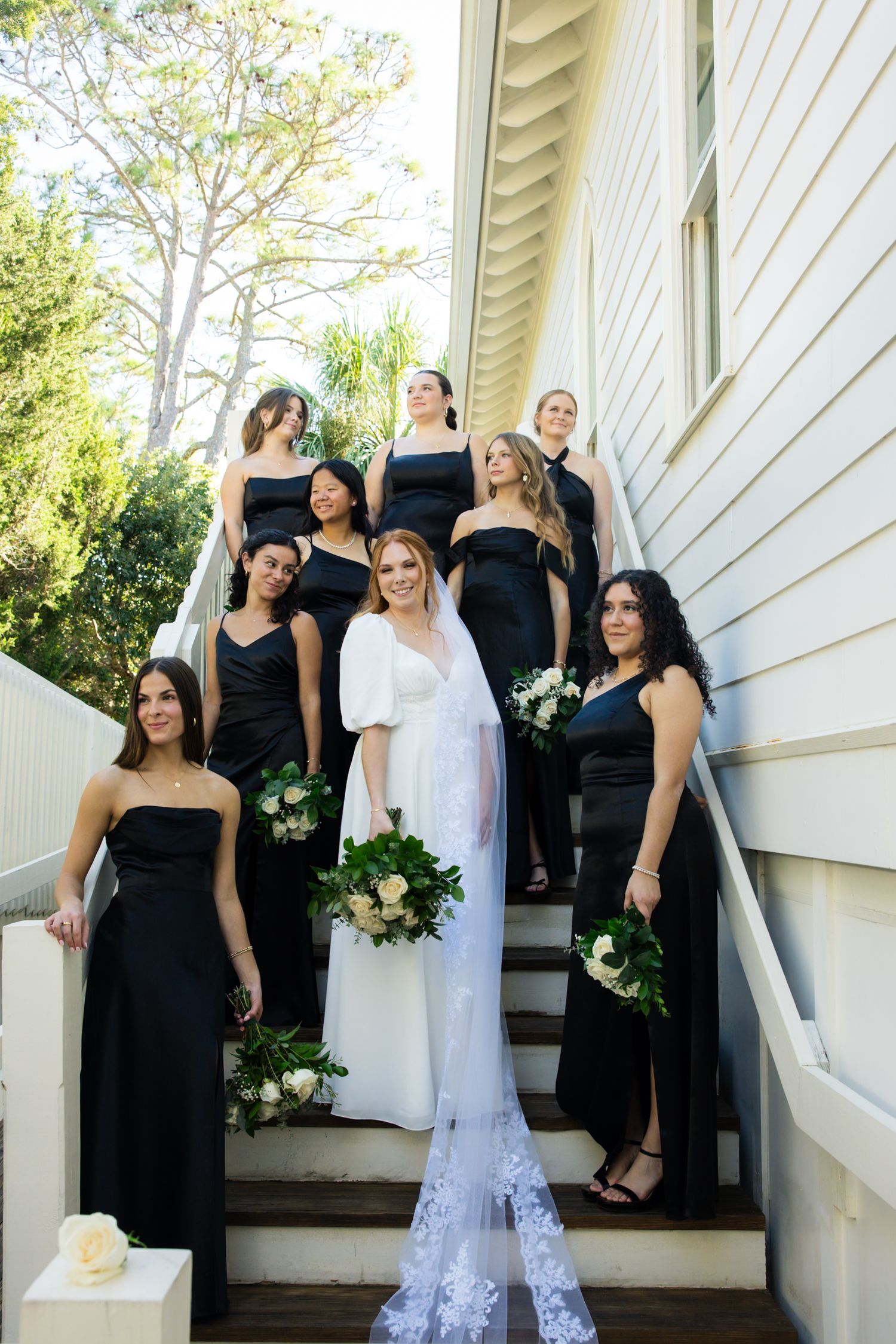 Wedding party in black dresses poses casually on white porch steps during outdoor celebration.