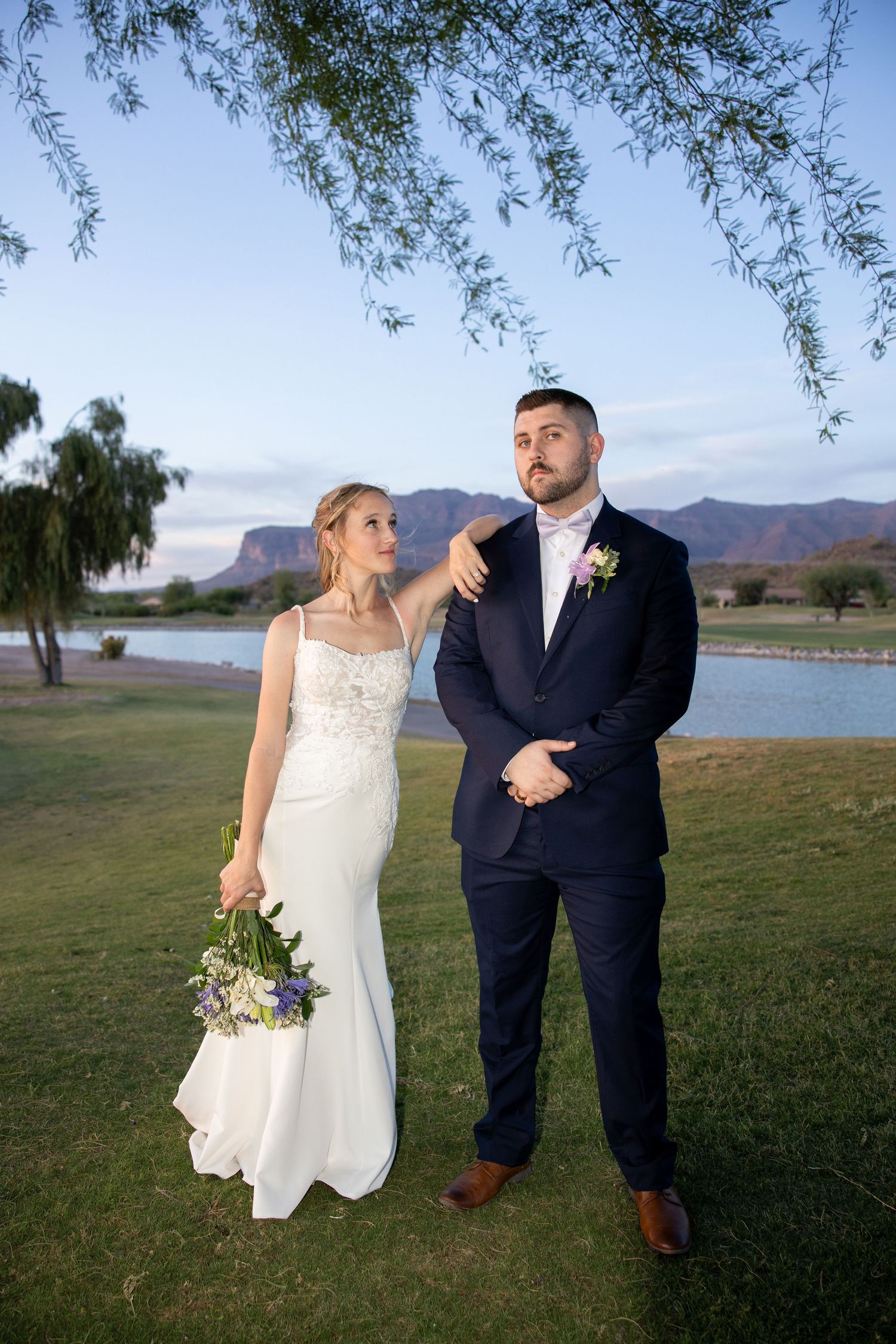 Bride and groom posing under willow tree with mountain lake backdrop at dusk.