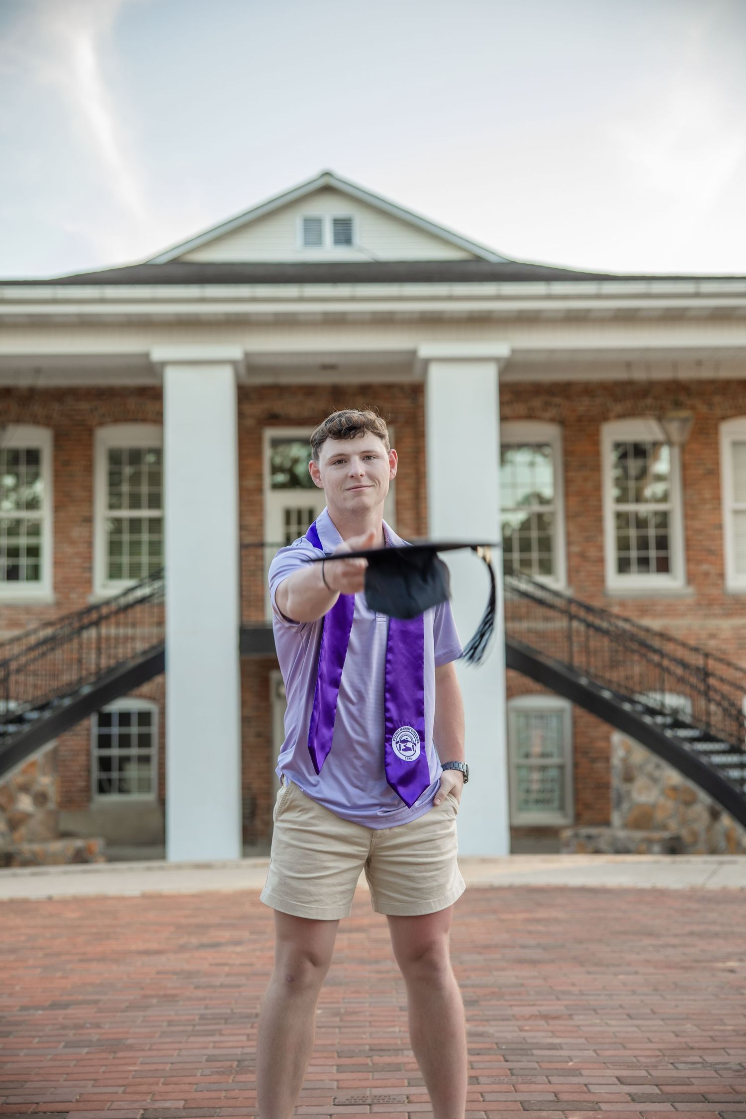 Graduate in purple stole poses with cap in front of brick university building.