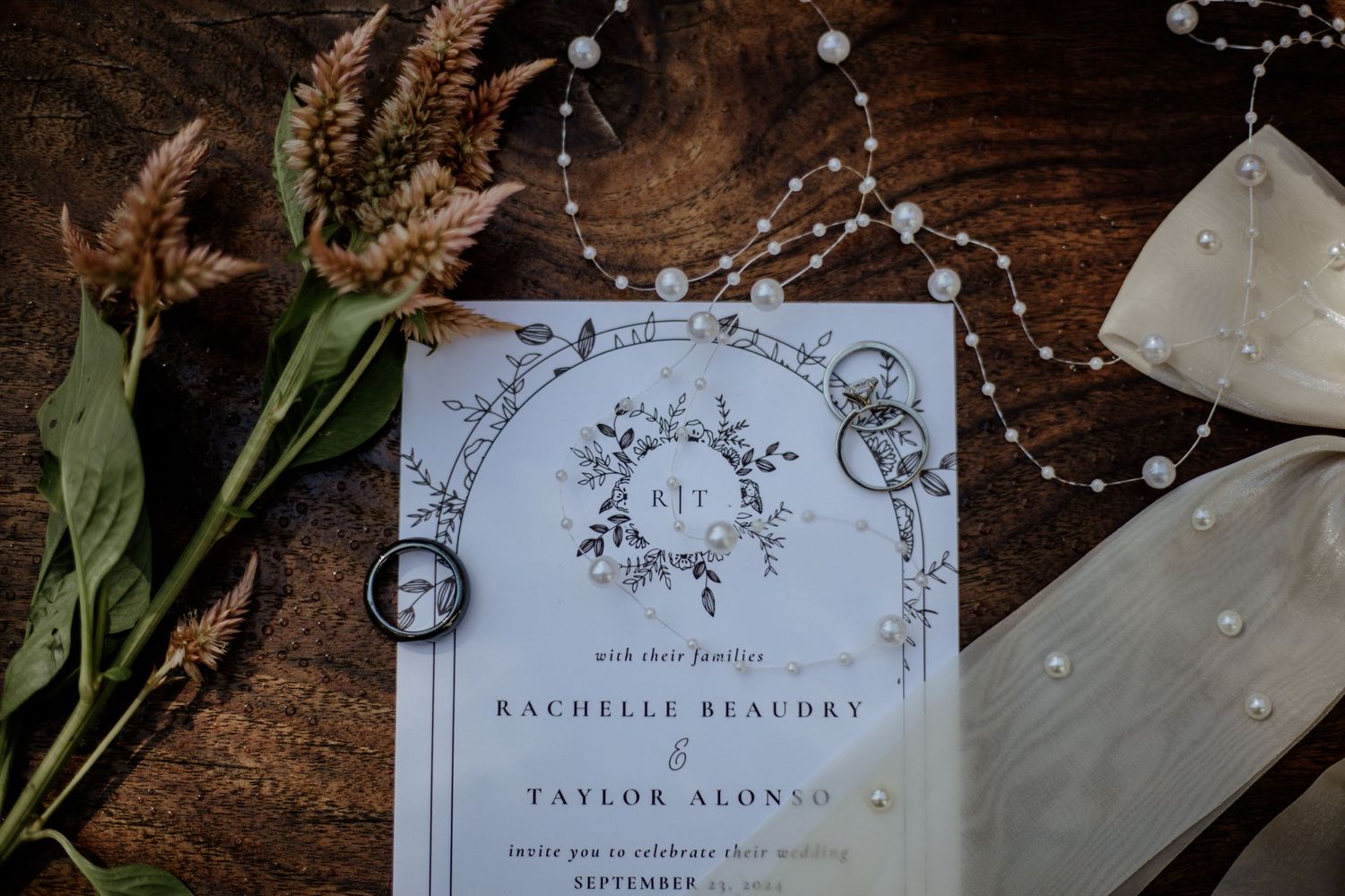 Wedding invitation with floral design lies on wooden surface with dried flowers and pearls.