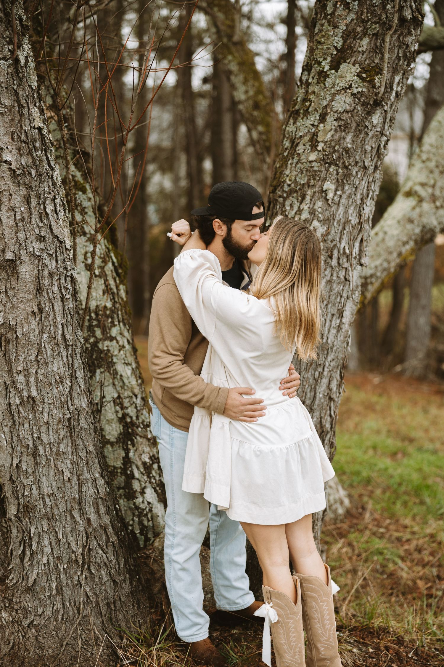 A romantic embrace between two people in casual attire against large tree trunks in a forest setting.