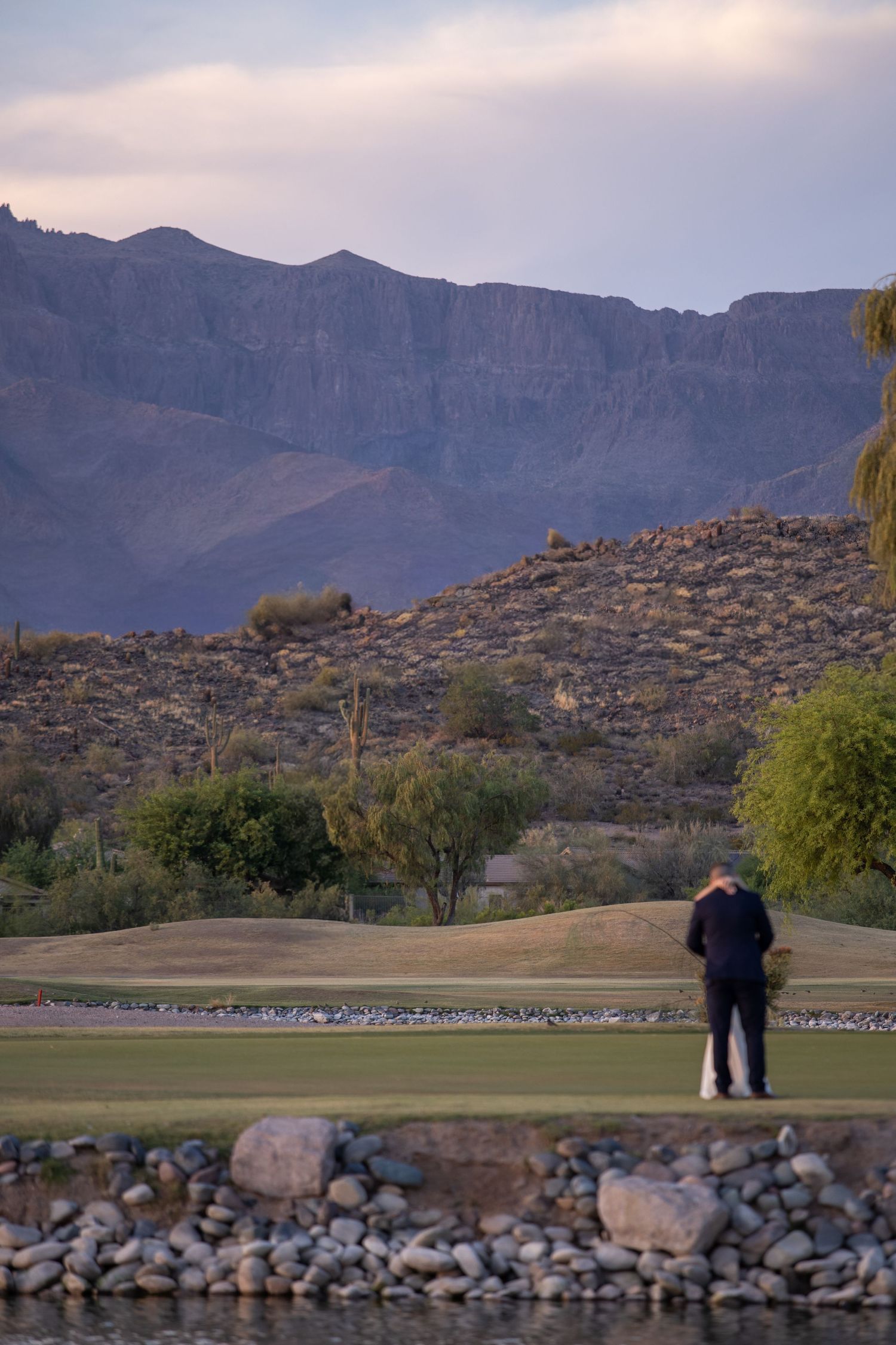 Lone figure standing on golf course green with dramatic rocky mountain landscape at dusk.