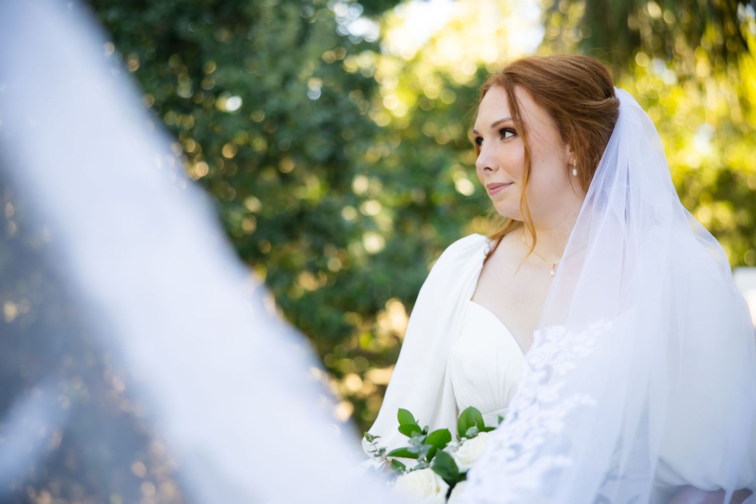 A bride in a white wedding gown stands outdoors surrounded by sunlit greenery and trees.
