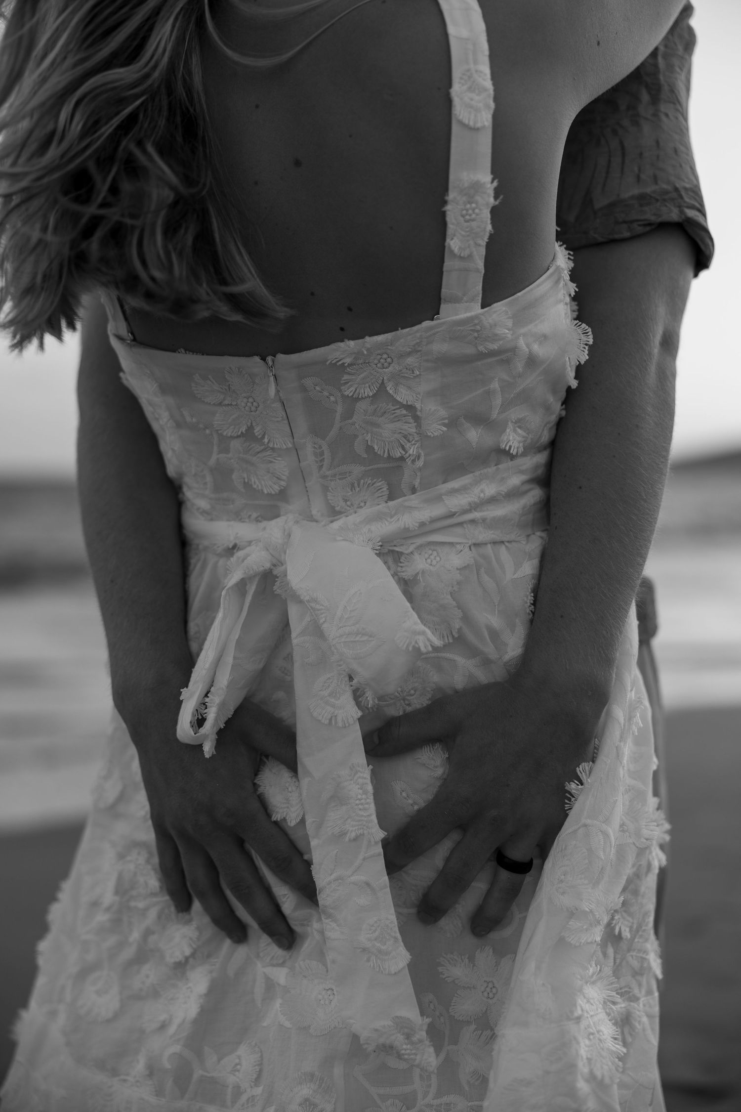 Black and white photo of hands resting on a textured white wedding dress with delicate bow detail.