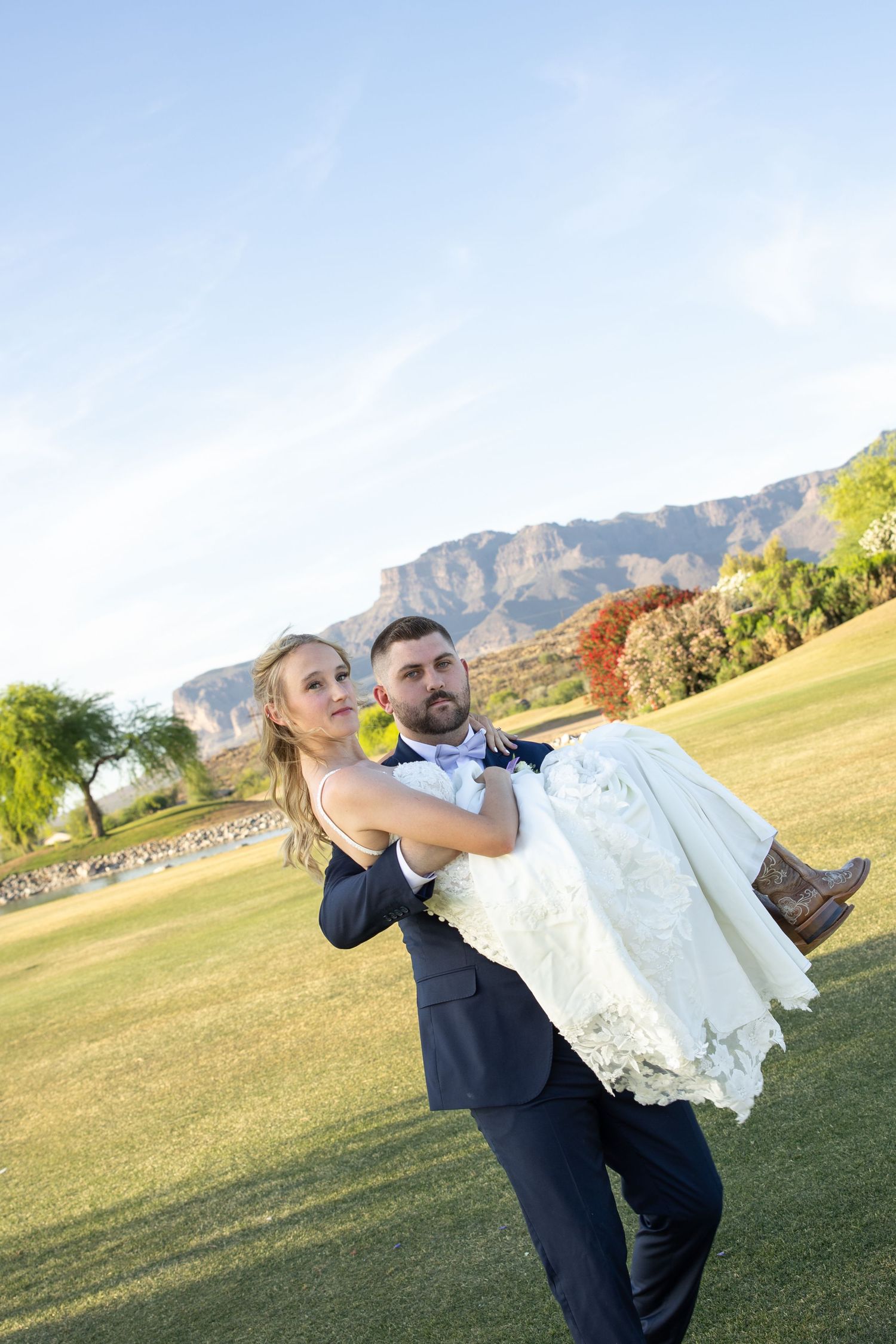 Groom carrying bride in white wedding dress on green lawn with mountain backdrop at golden hour.