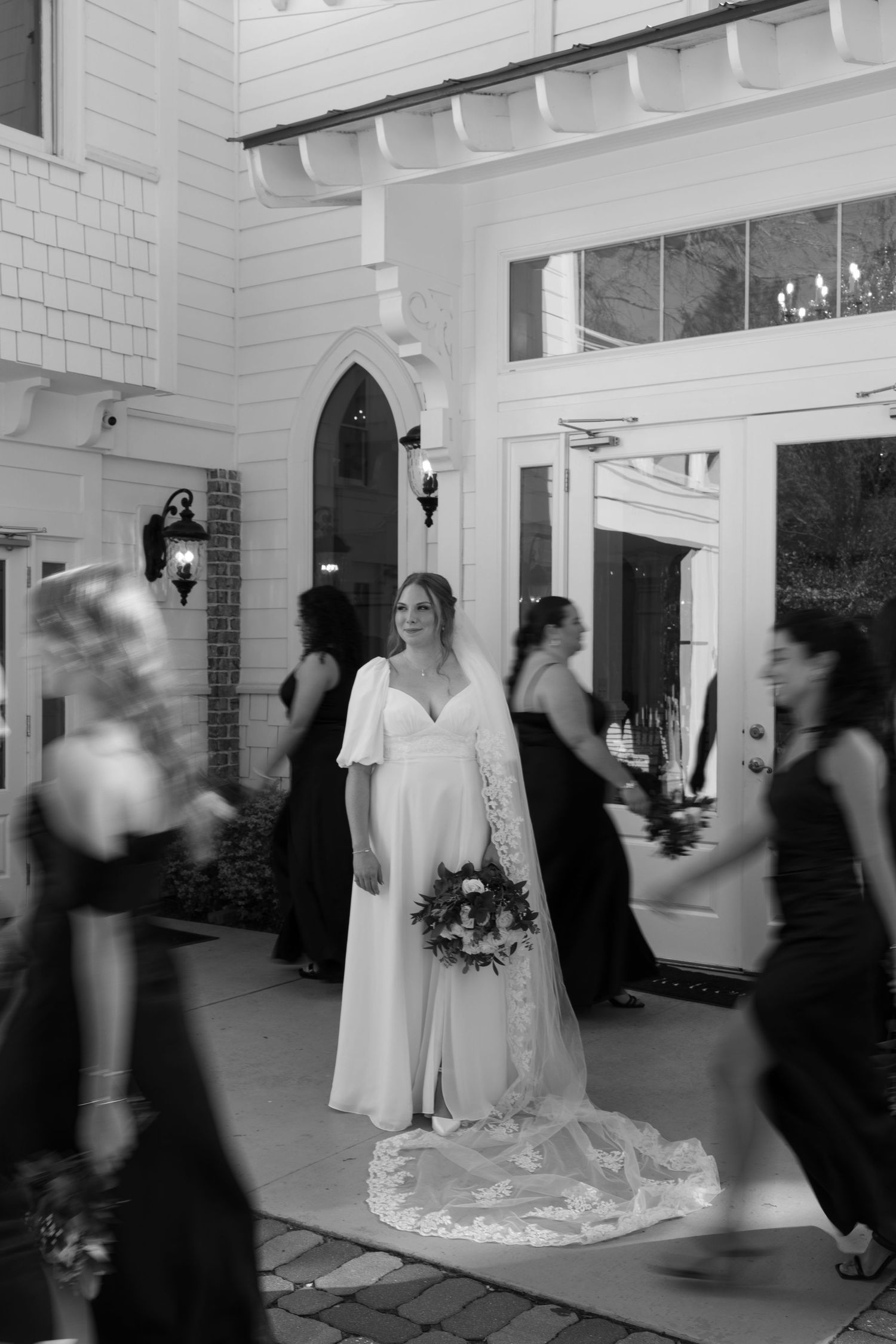 A black and white wedding photo showing guests mingling in a hallway with white brick walls and a bride in the center.