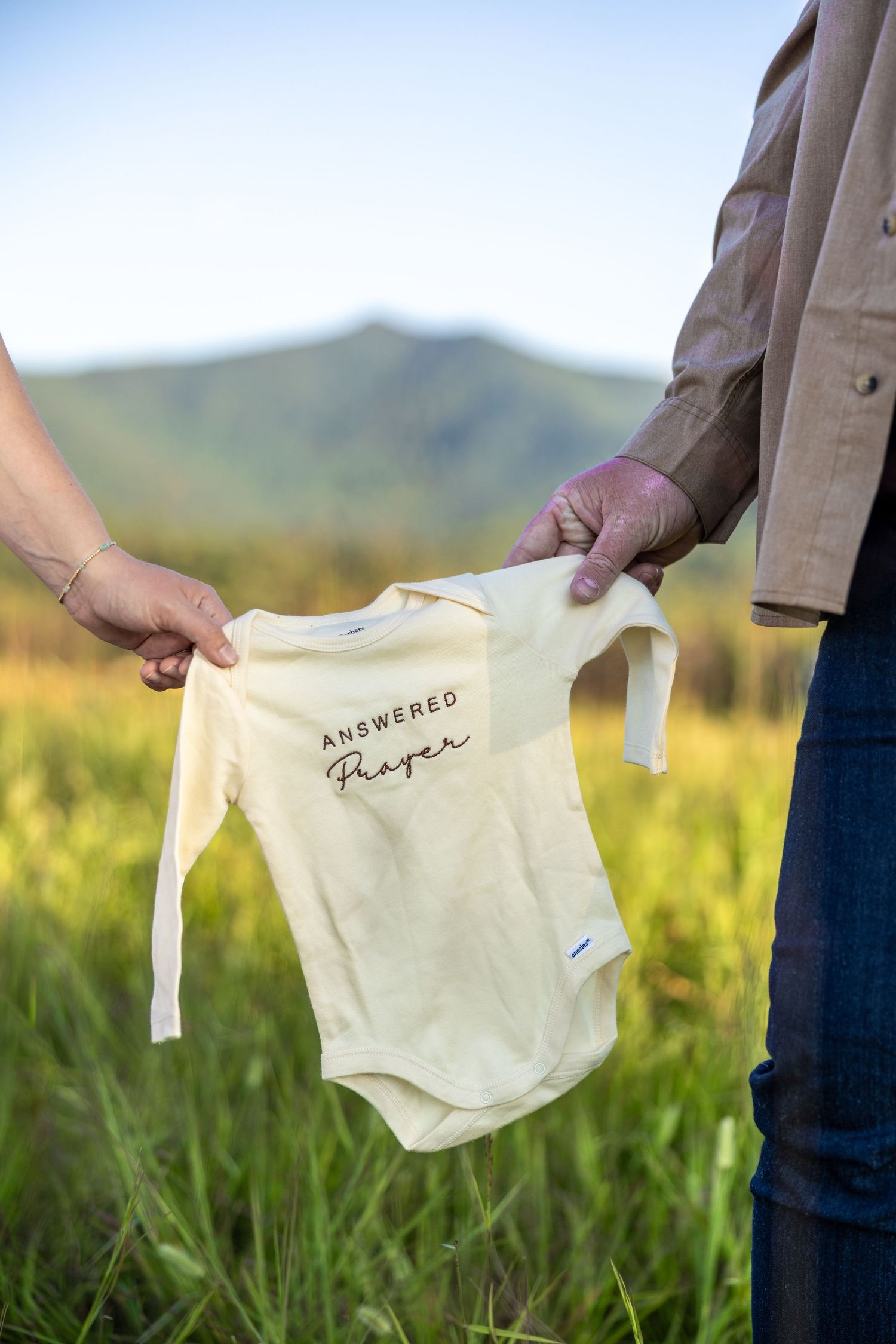 Multiple hands hold up small beige baby onesies with text embroidery against a blurred natural outdoor background.