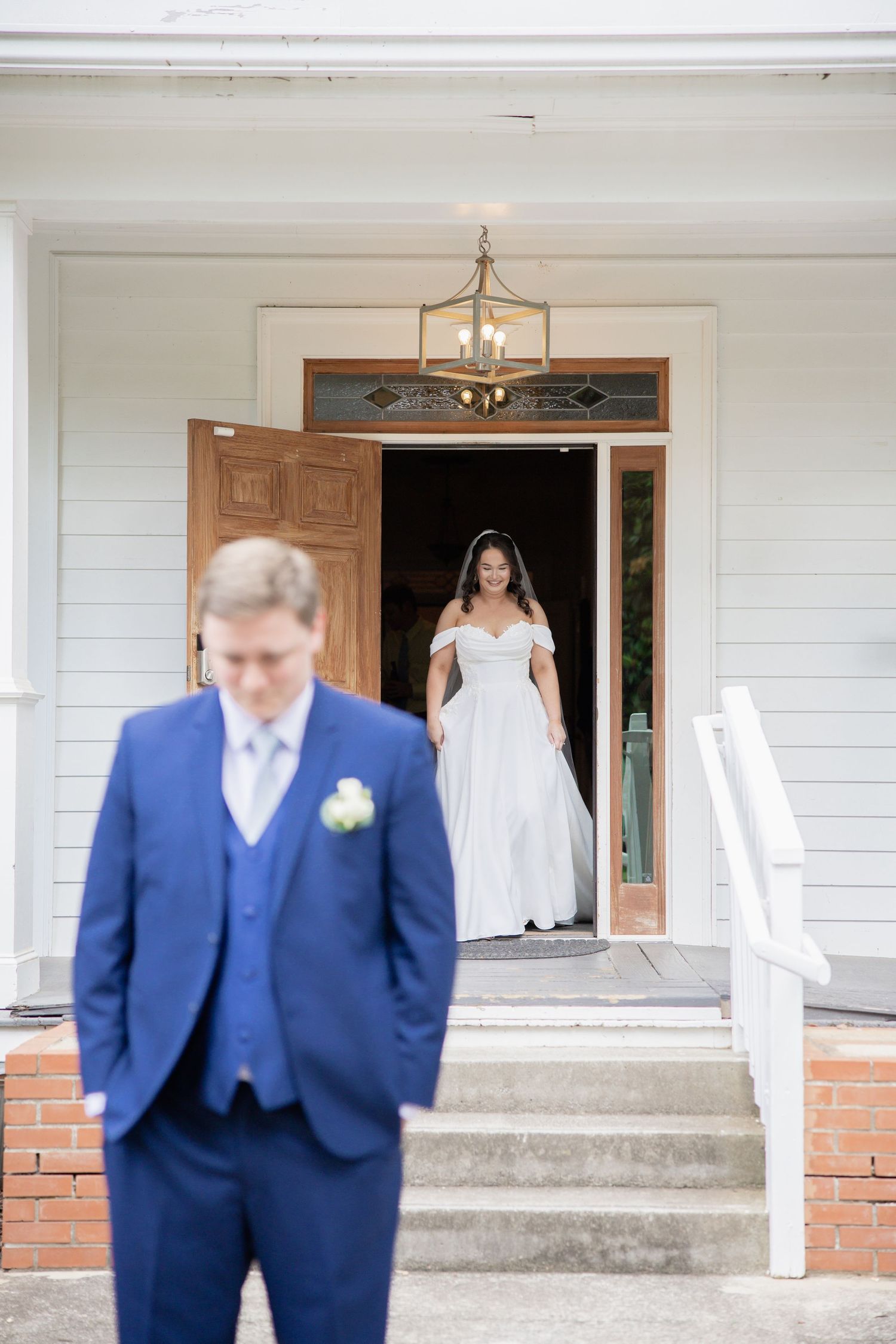 First look moment between bride in white dress and groom in navy suit on porch steps.