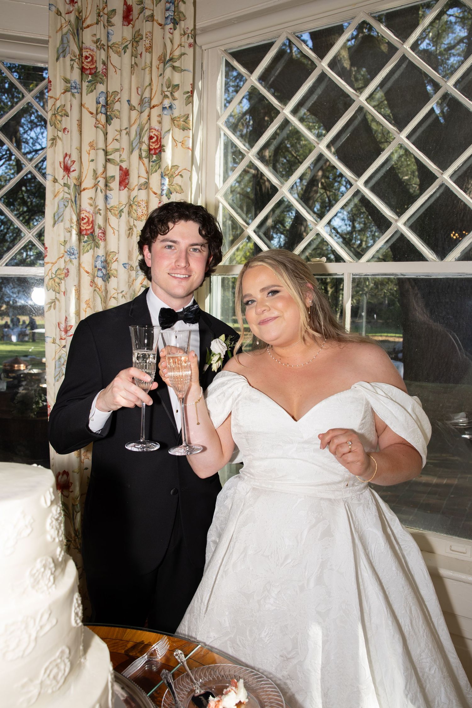 A newlywed couple toasts with champagne glasses at their wedding reception by a latticed window with floral curtains.