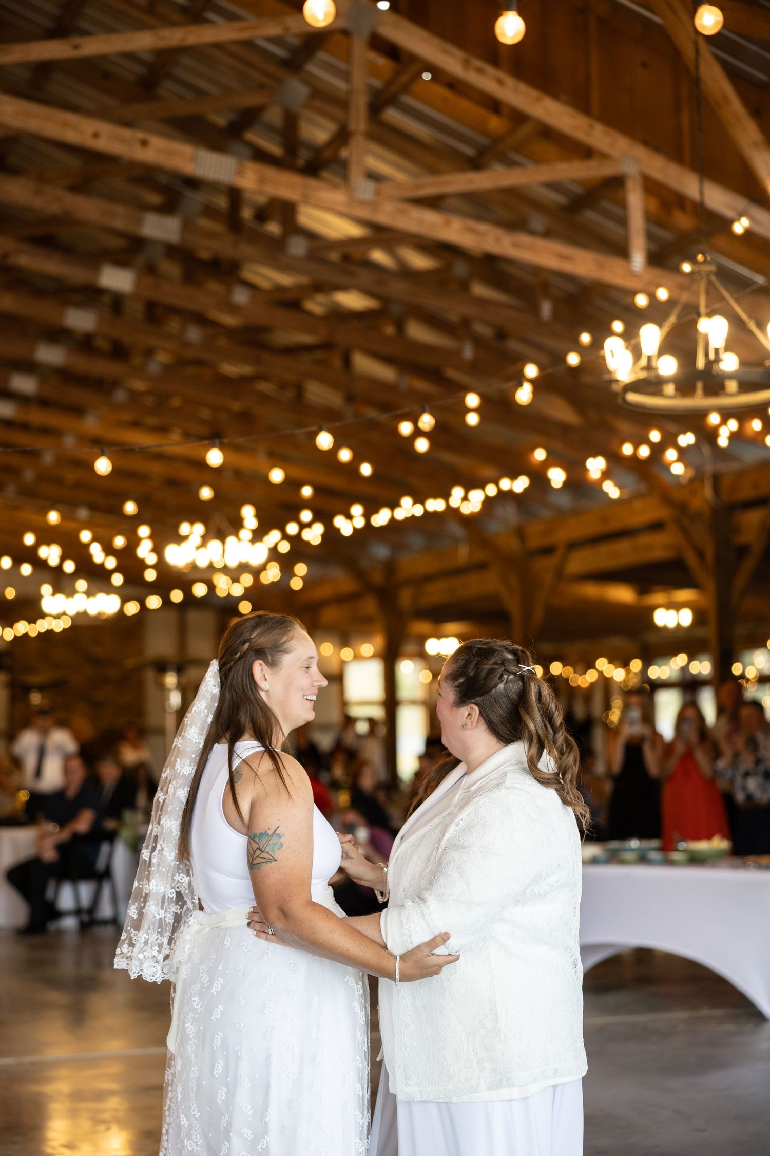 A couple shares their first dance as newlyweds in a rustic barn venue decorated with romantic string lights overhead.