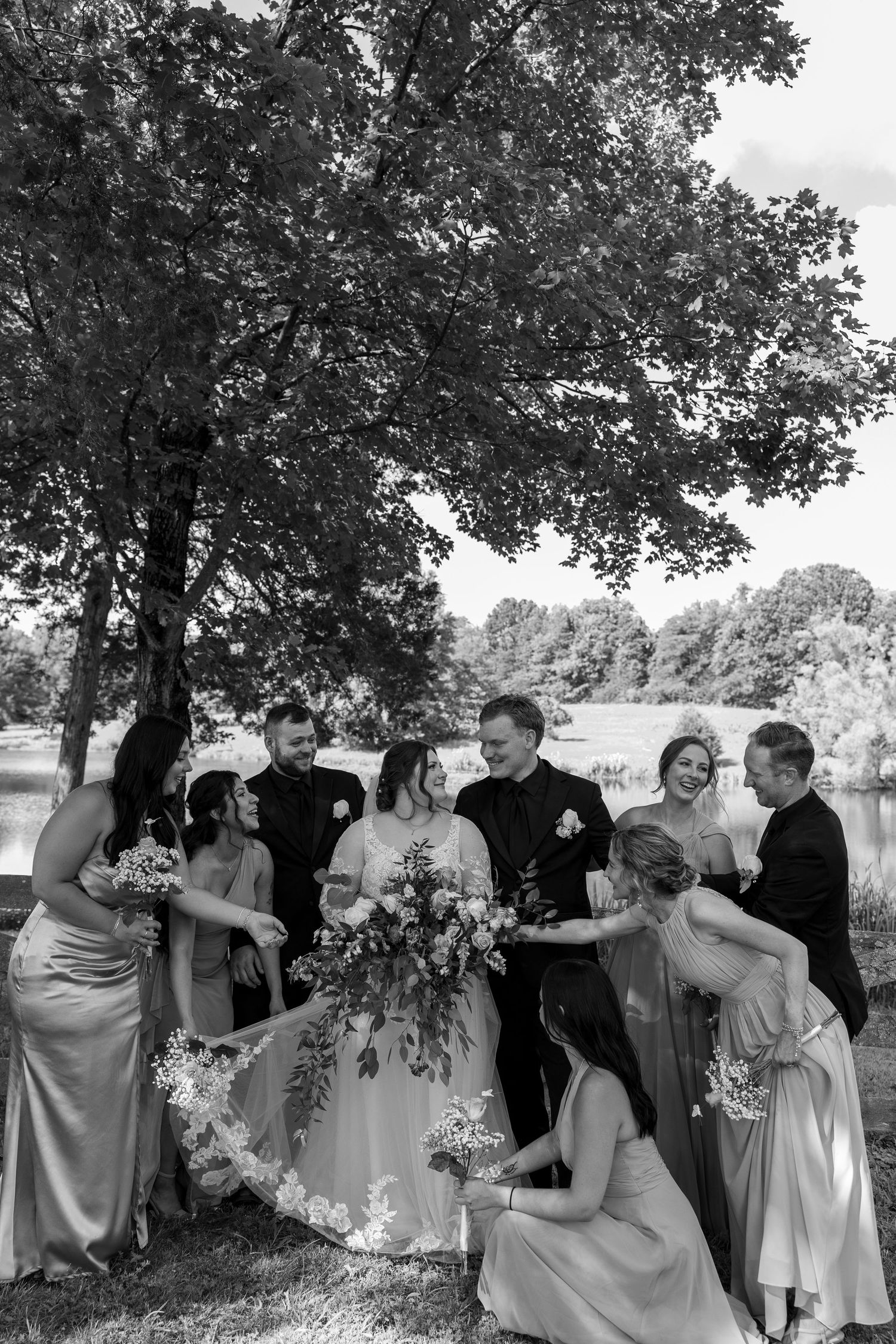 Black and white wedding party photo under a large tree, bride holding bouquet with veil being arranged by attendants.