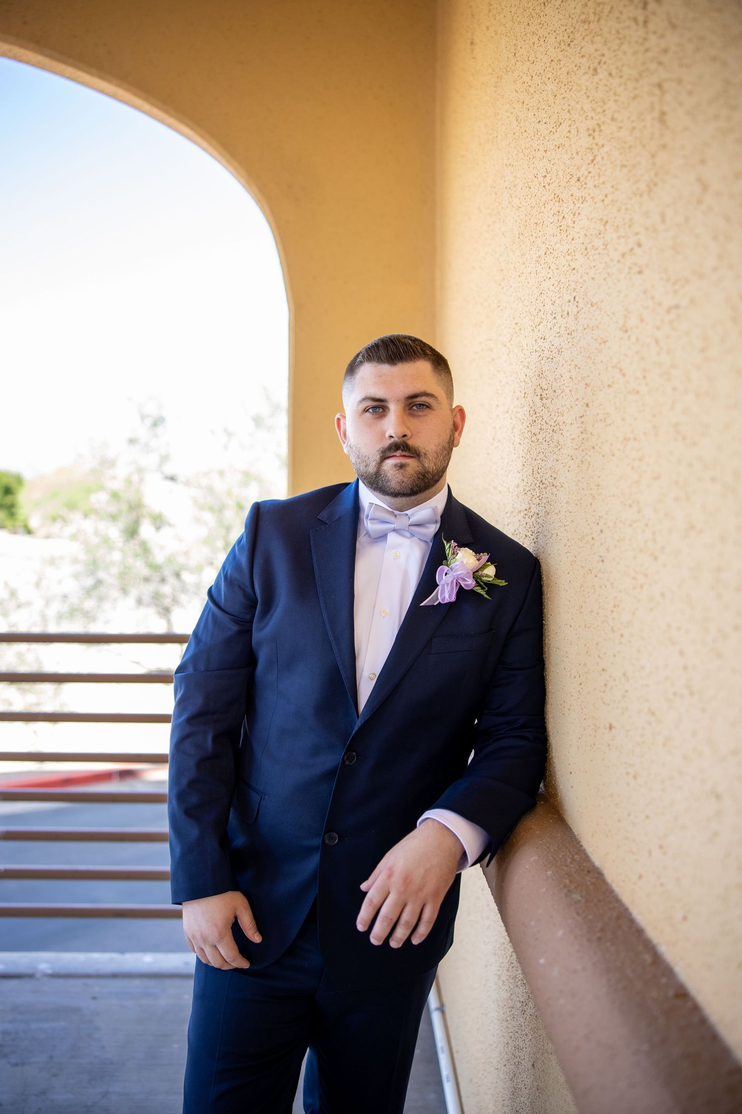 Groom in navy suit with bow tie and boutonniere leaning against wall at outdoor wedding venue.