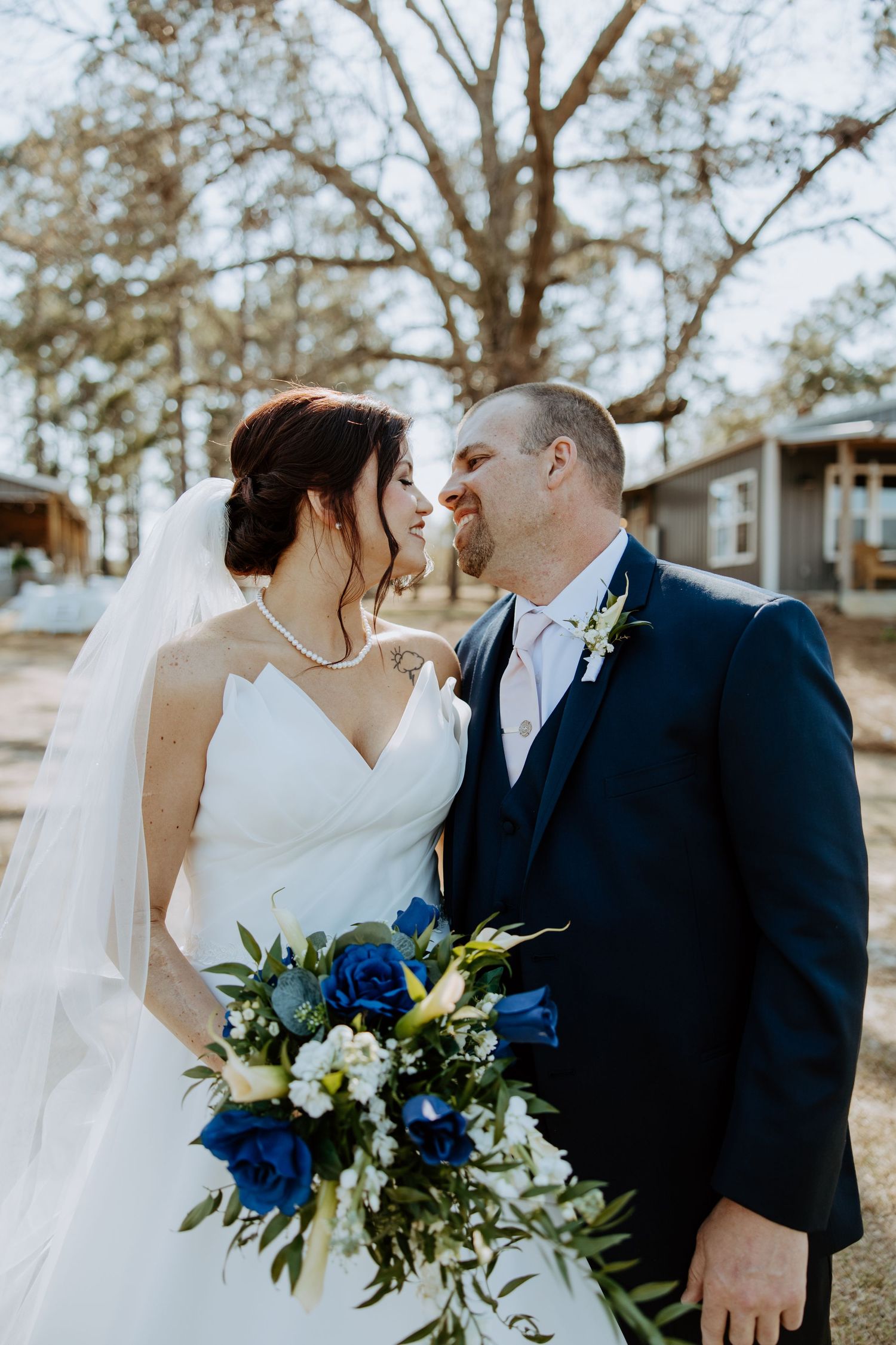 A wedding portrait showing a couple in formal attire with a blue and white bouquet against a natural backdrop.