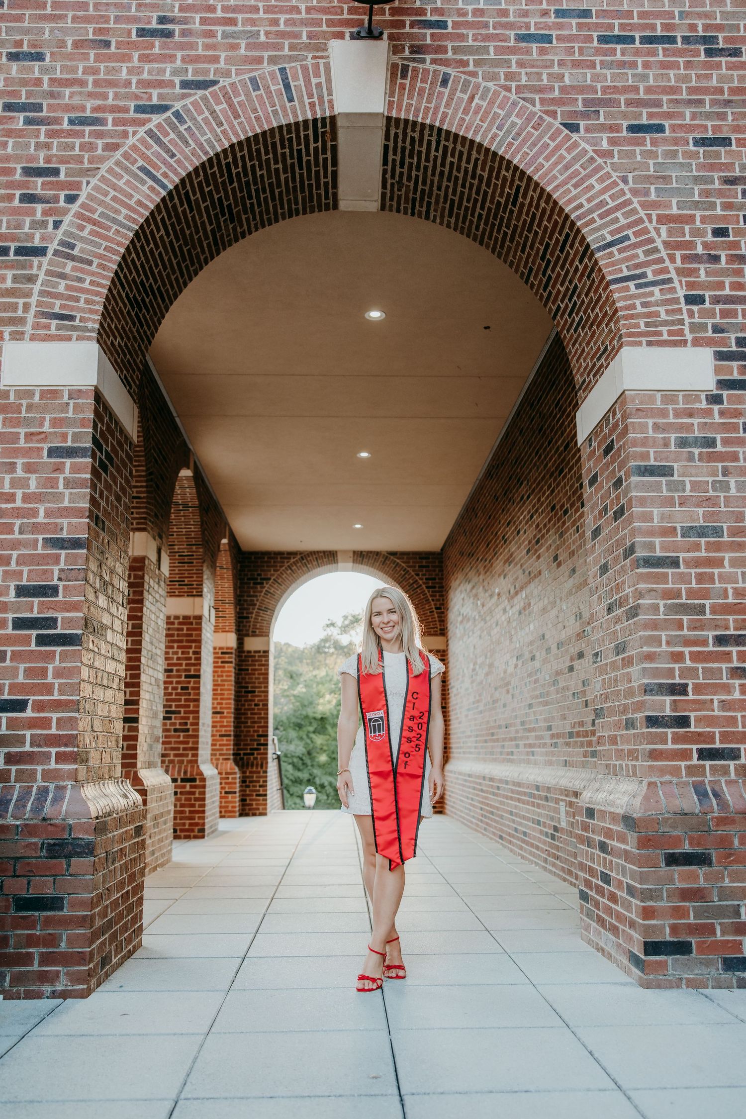 Graduate walks through arched brick hallway wearing red stole and matching heels.