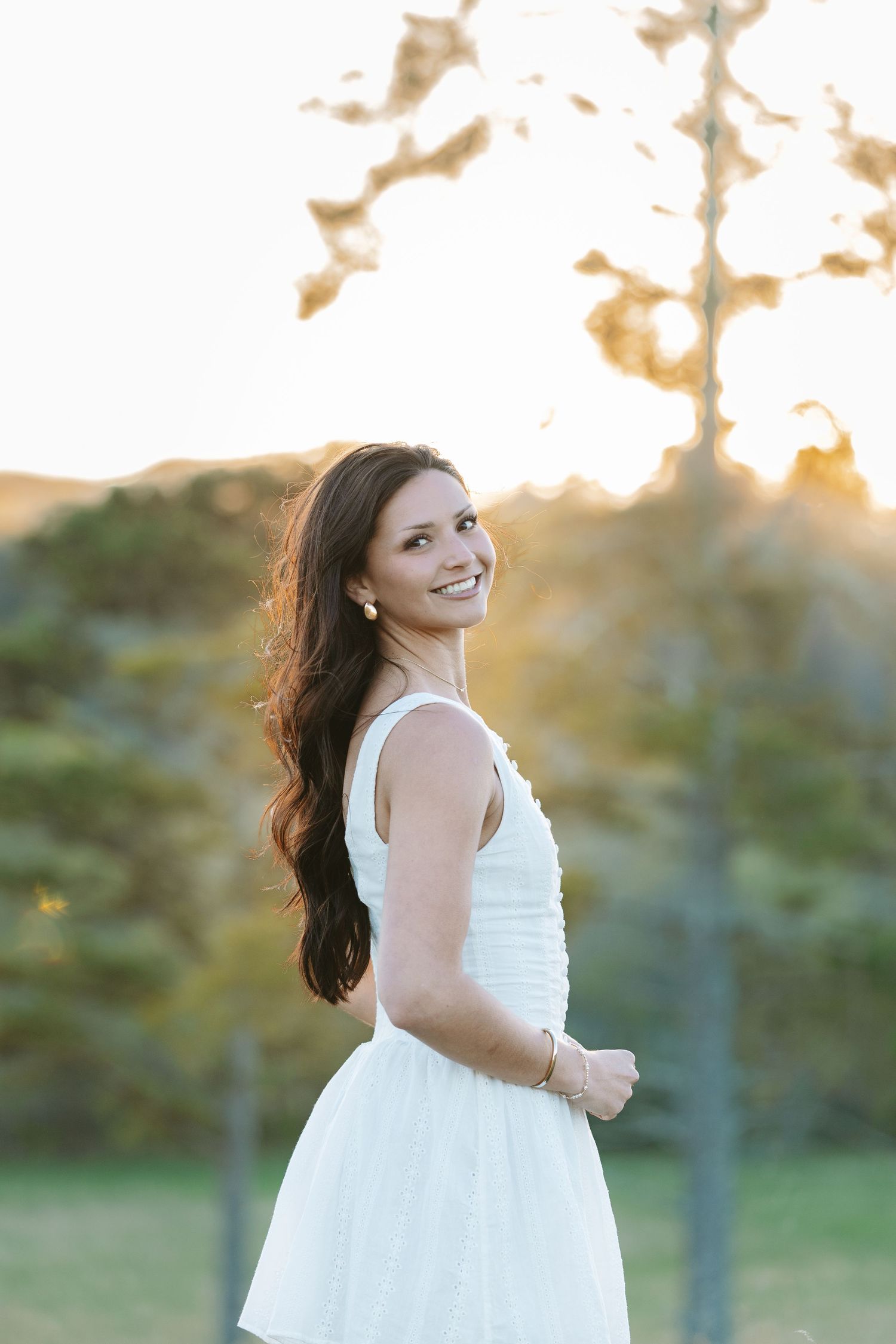 Person in white dress poses outdoors during sunset with trees in background.