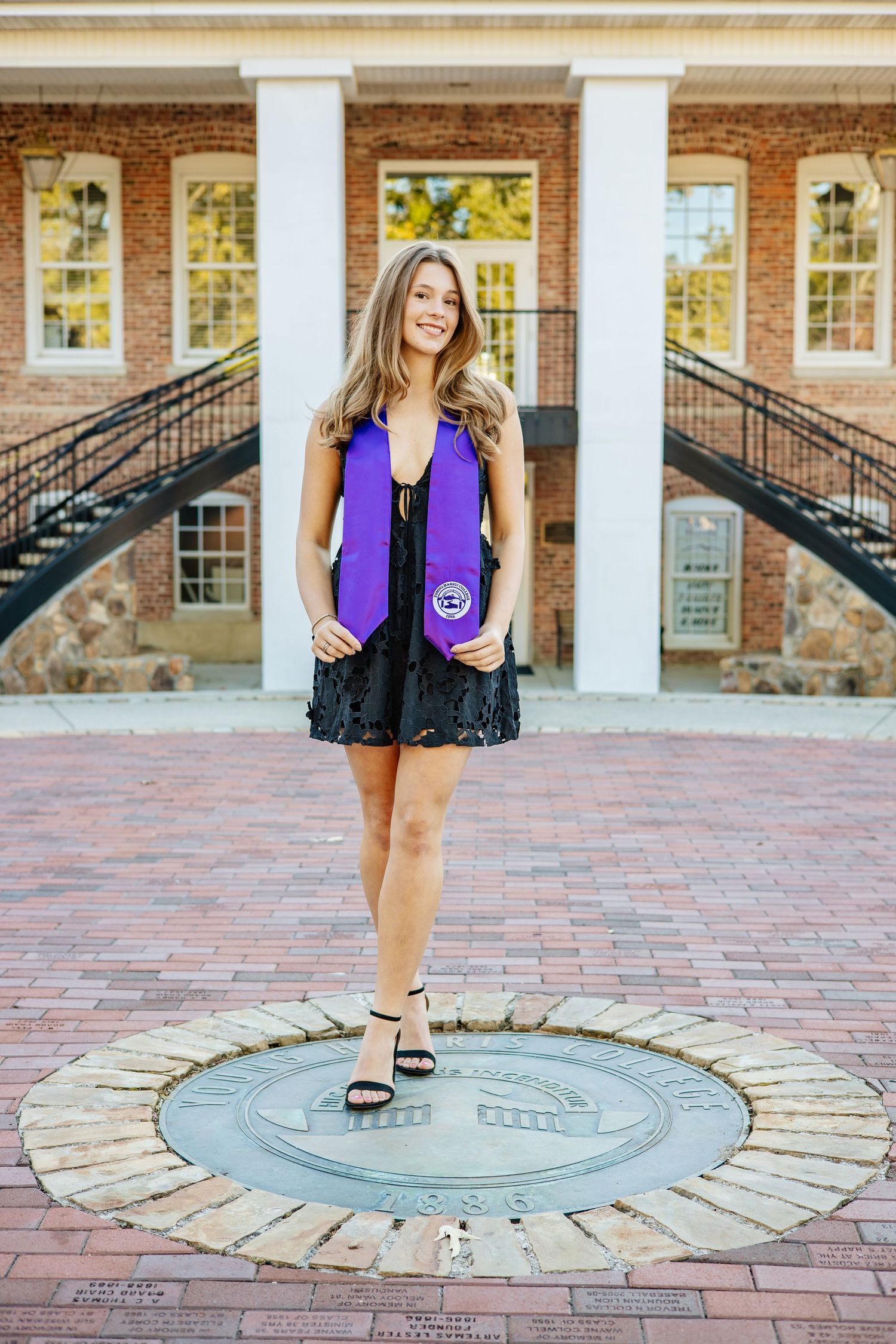 Graduate in purple stole and black dress stands on circular brick pattern in front of campus building.