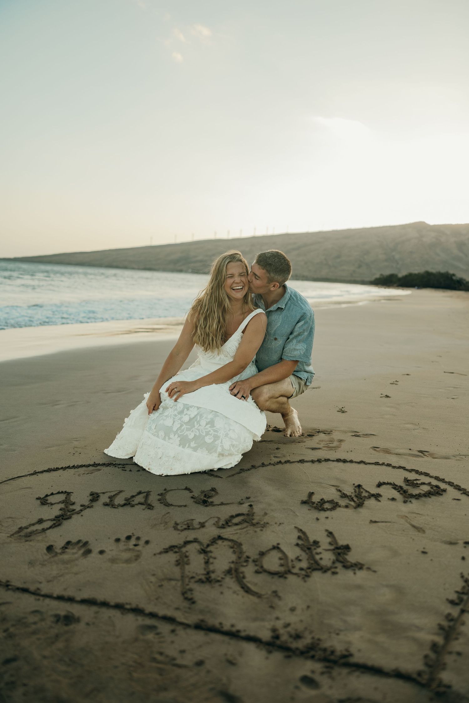 A couple shares a romantic moment at the beach near text written in the sand during sunset.