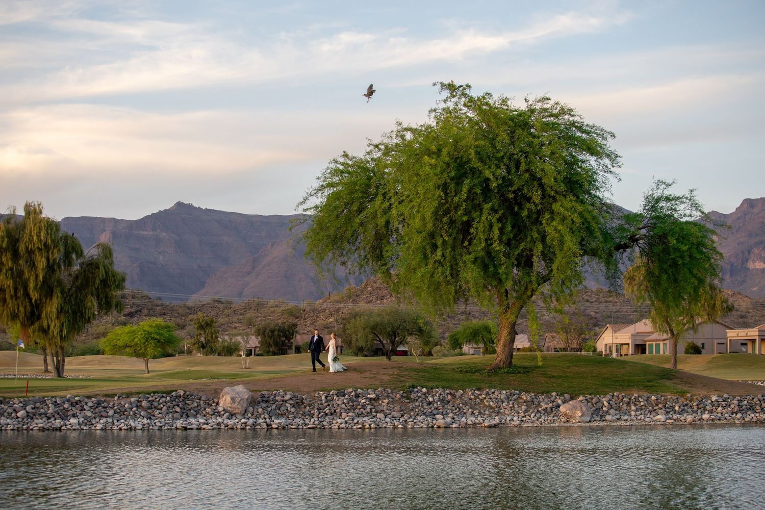 Bride and groom walking near a lake with willow trees and mountain views at sunset.