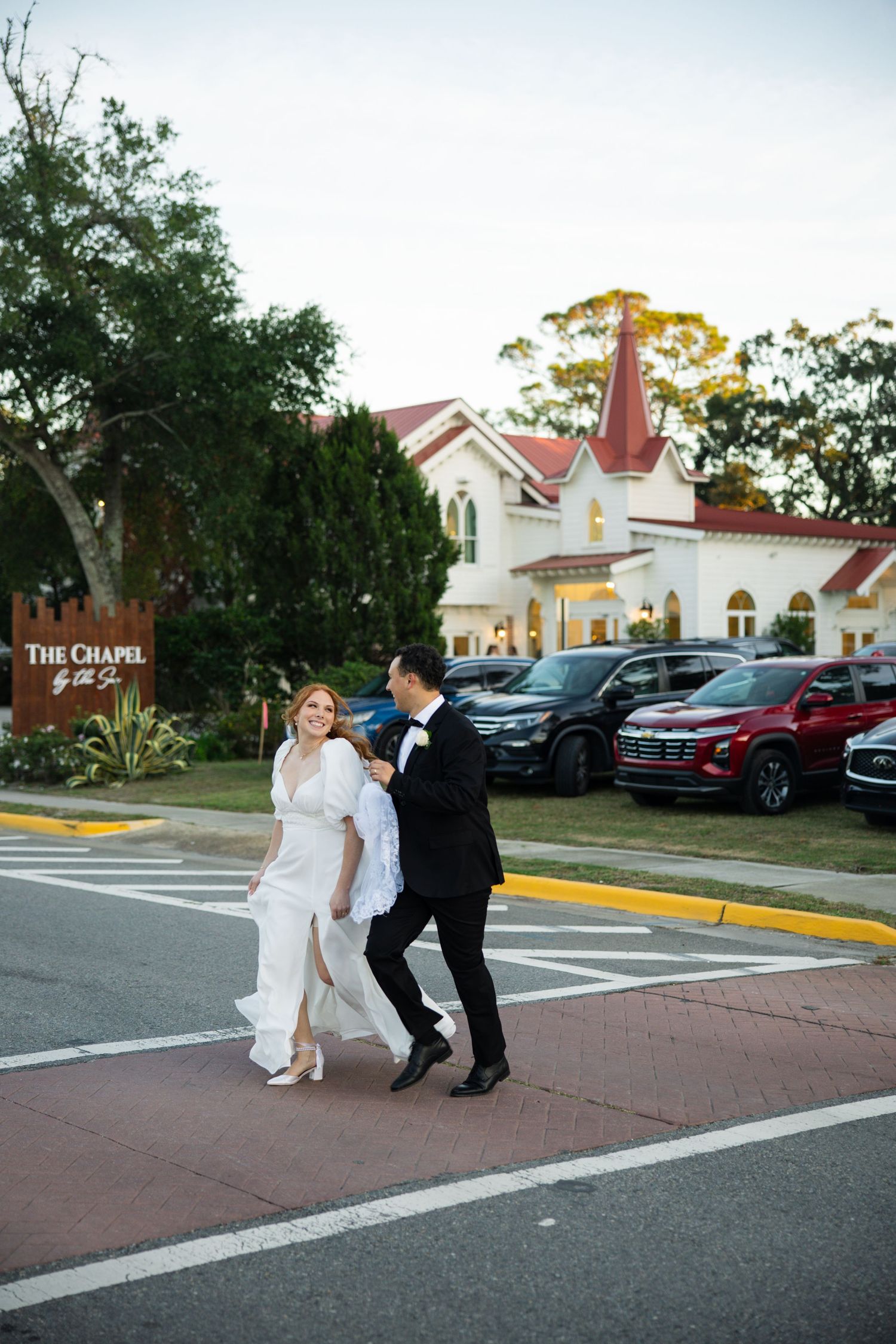 A couple in wedding attire playfully walks through a parking lot with a white chapel visible in the background.