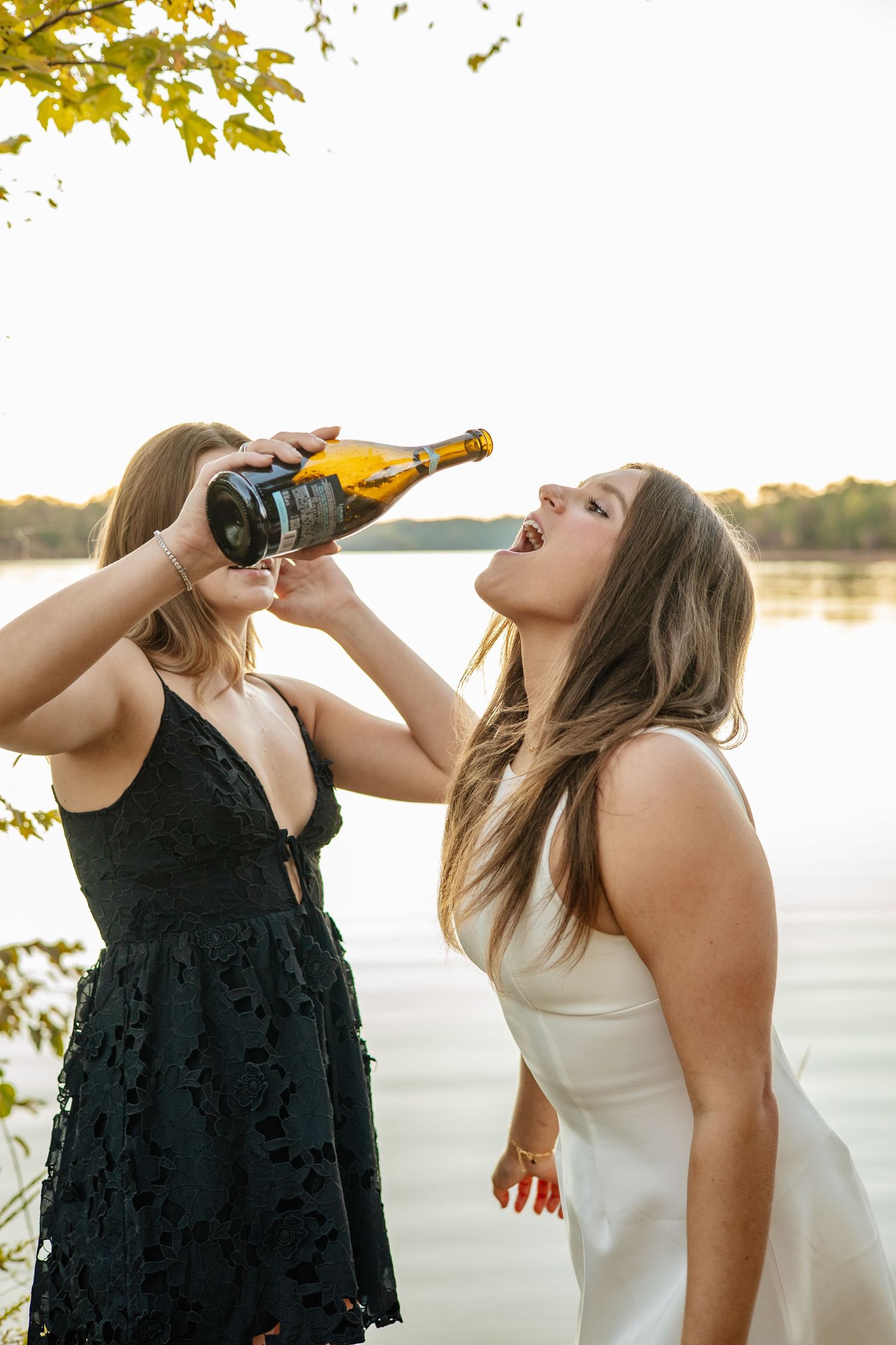 Two friends in formal dresses drinking champagne by lakeside at sunset.