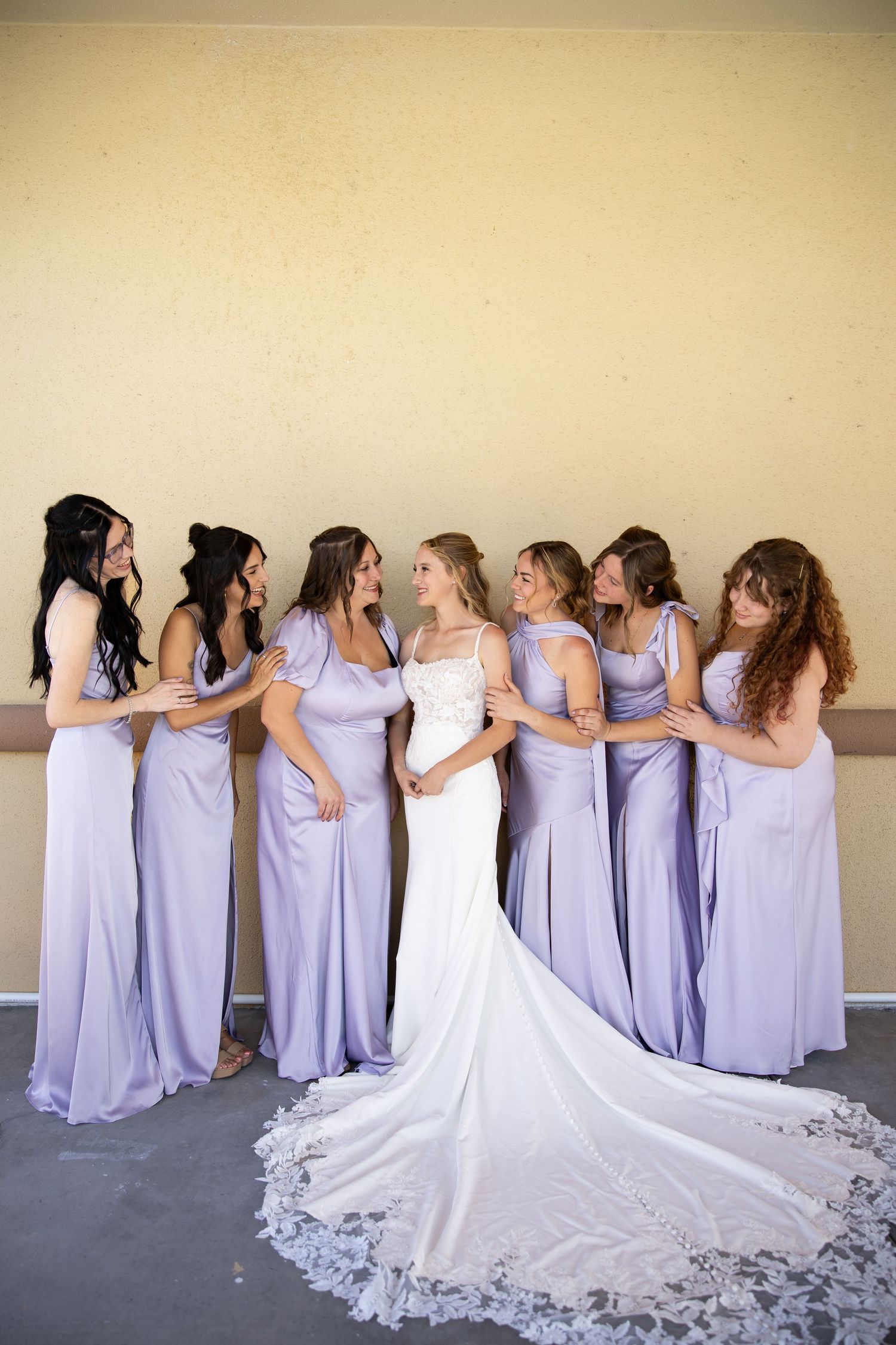 Bride in white lace gown surrounded by six bridesmaids in lavender dresses smiling together indoors.