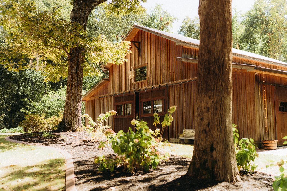 A rustic wooden barn-style house nestled among tall trees with dappled sunlight filtering through the branches.
