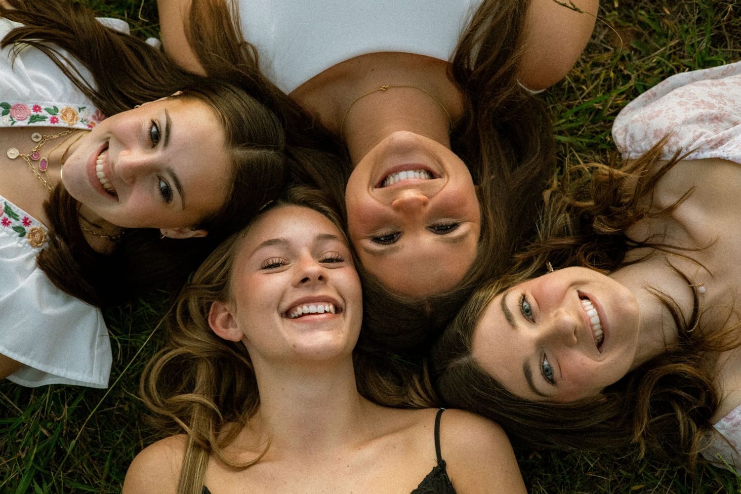 Four friends laying on grass with their heads together forming a circle and smiling.