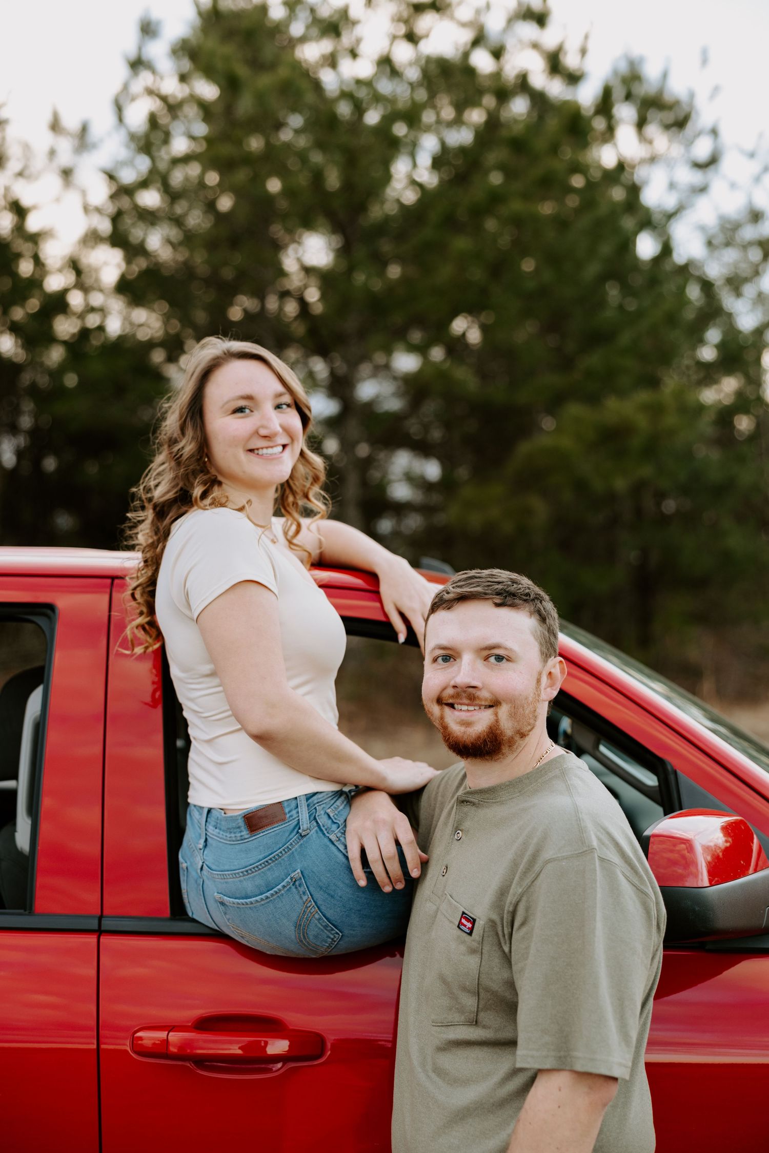 A couple poses playfully by a red pickup truck during a sunset photoshoot.