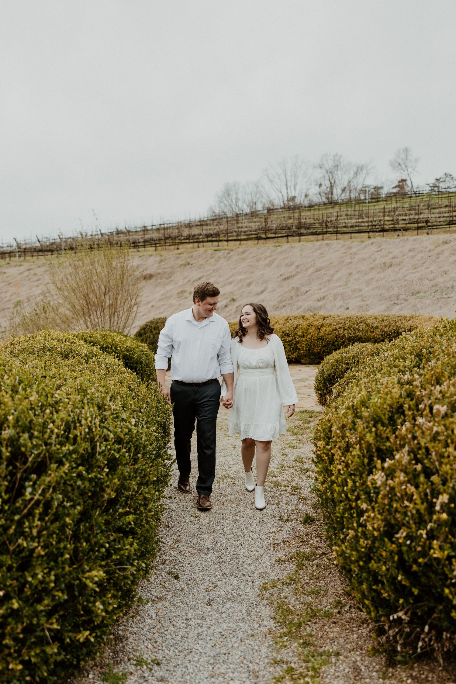 A couple walking together on a gravel path lined with manicured hedges on an overcast day.