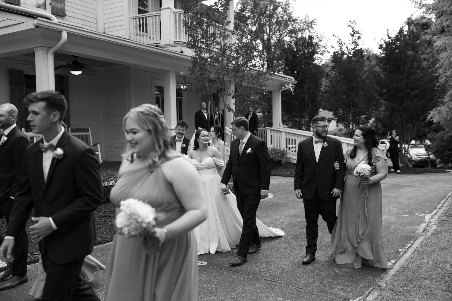 Wedding party members in formal attire walk outside a white columned building.