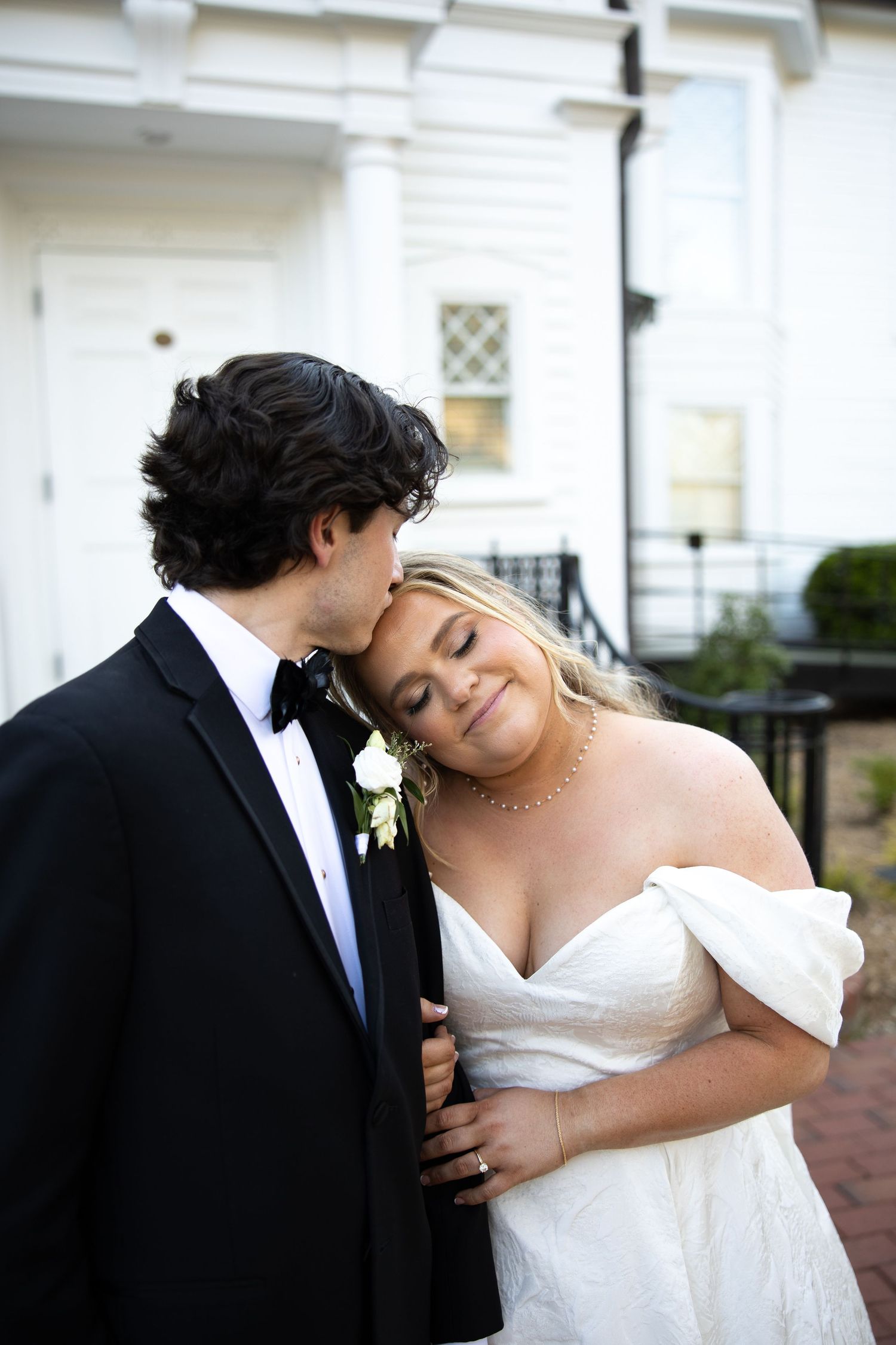 Groom in black tuxedo kissing bride in white off-shoulder gown outside elegant white building.