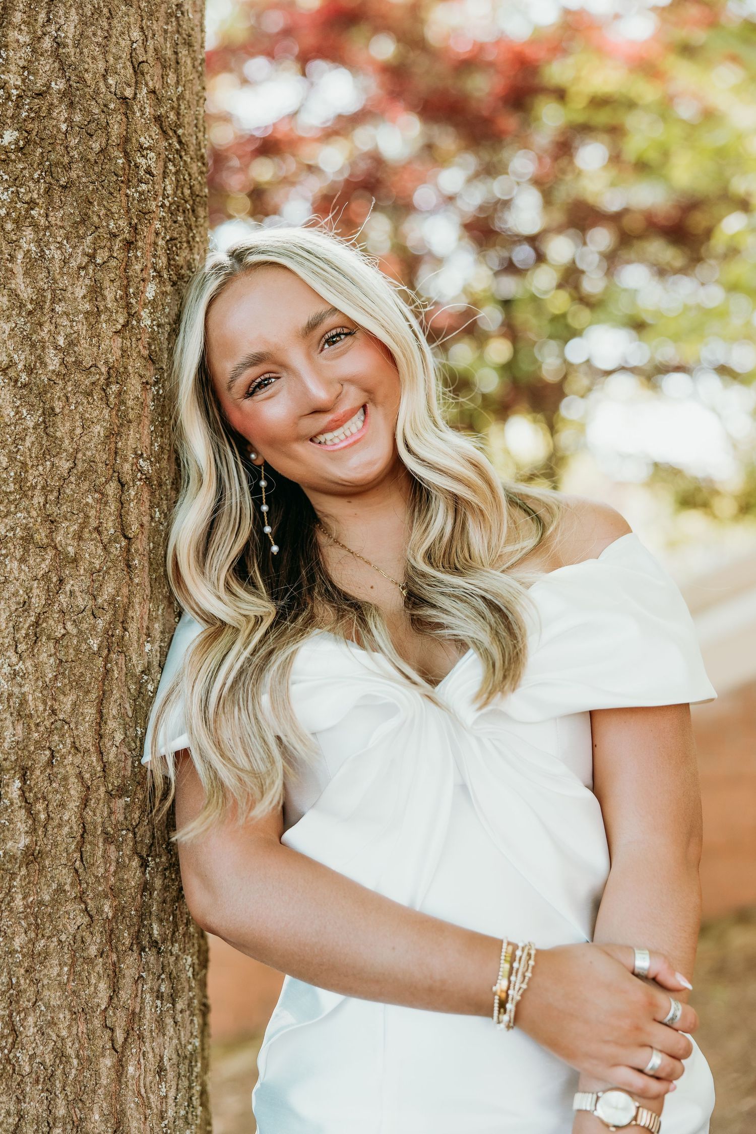 Portrait of person in white dress leaning against tree with autumn foliage.