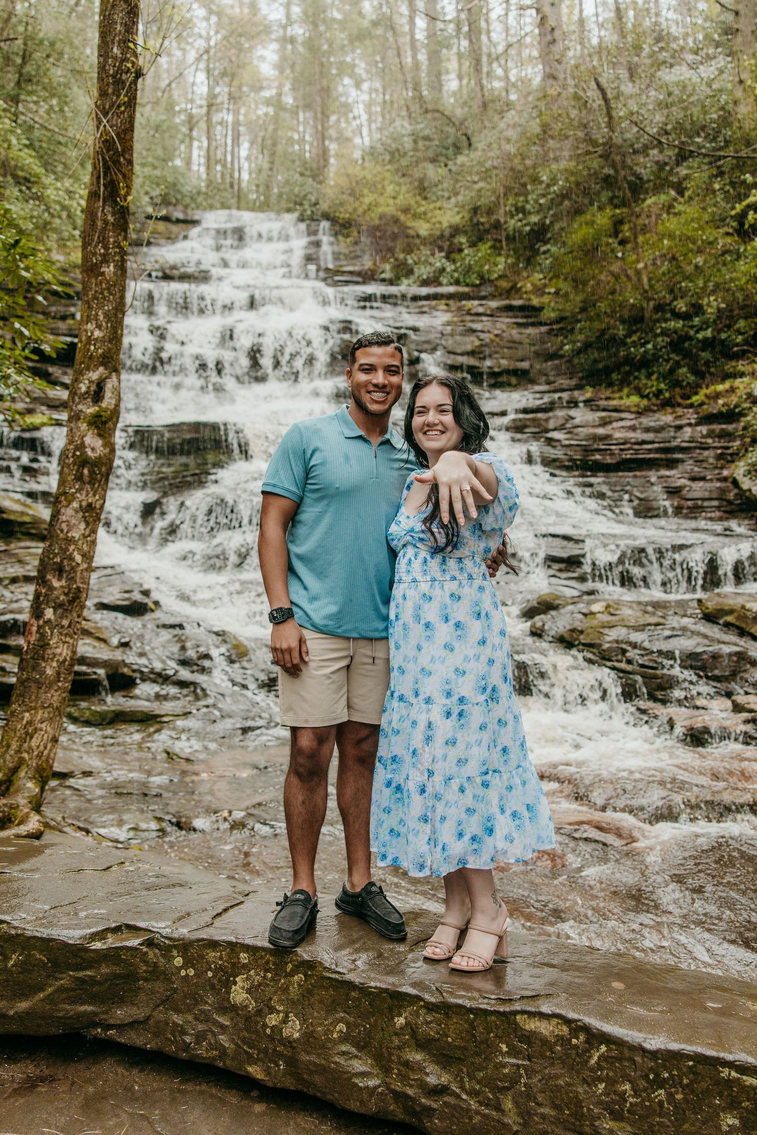 A couple poses together in front of a cascading waterfall wearing coordinated blue and khaki outfits.