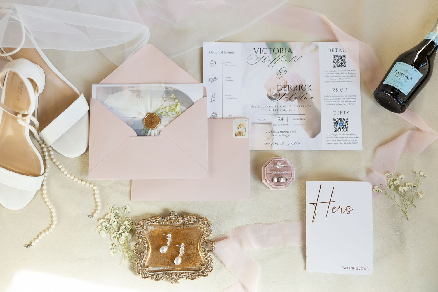 Elegant wedding details including pink invitation suite, white heels, pearl necklace, and wine bottle arranged on light surface.