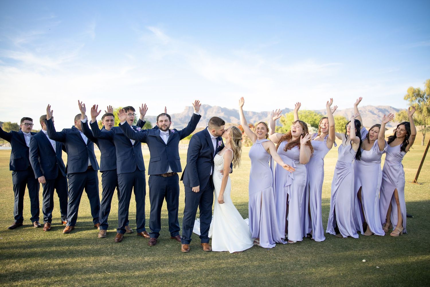 Wedding party celebrating outdoors, bridesmaids in lavender gowns and groomsmen in navy suits cheering with arms raised.