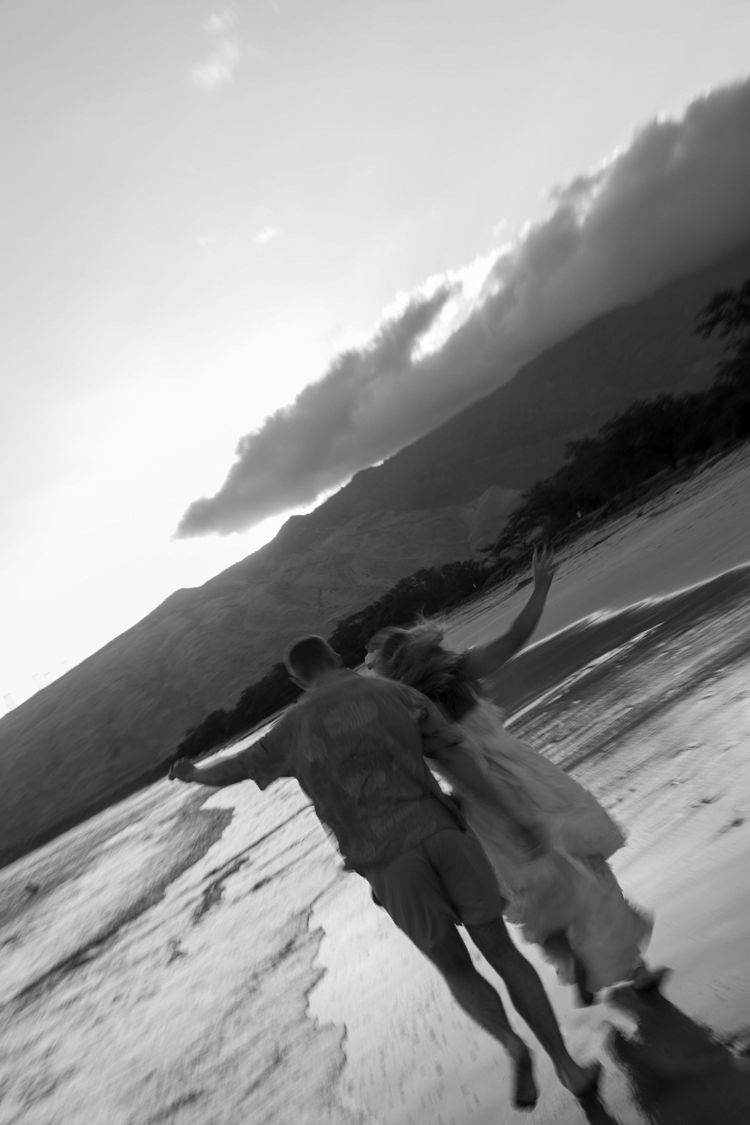 Two people running together on a black and white beach with dramatic clouds and mountains in the background.
