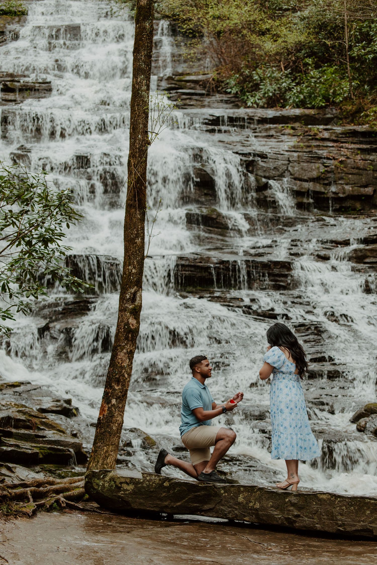 A romantic proposal scene in front of a cascading waterfall with one person kneeling.