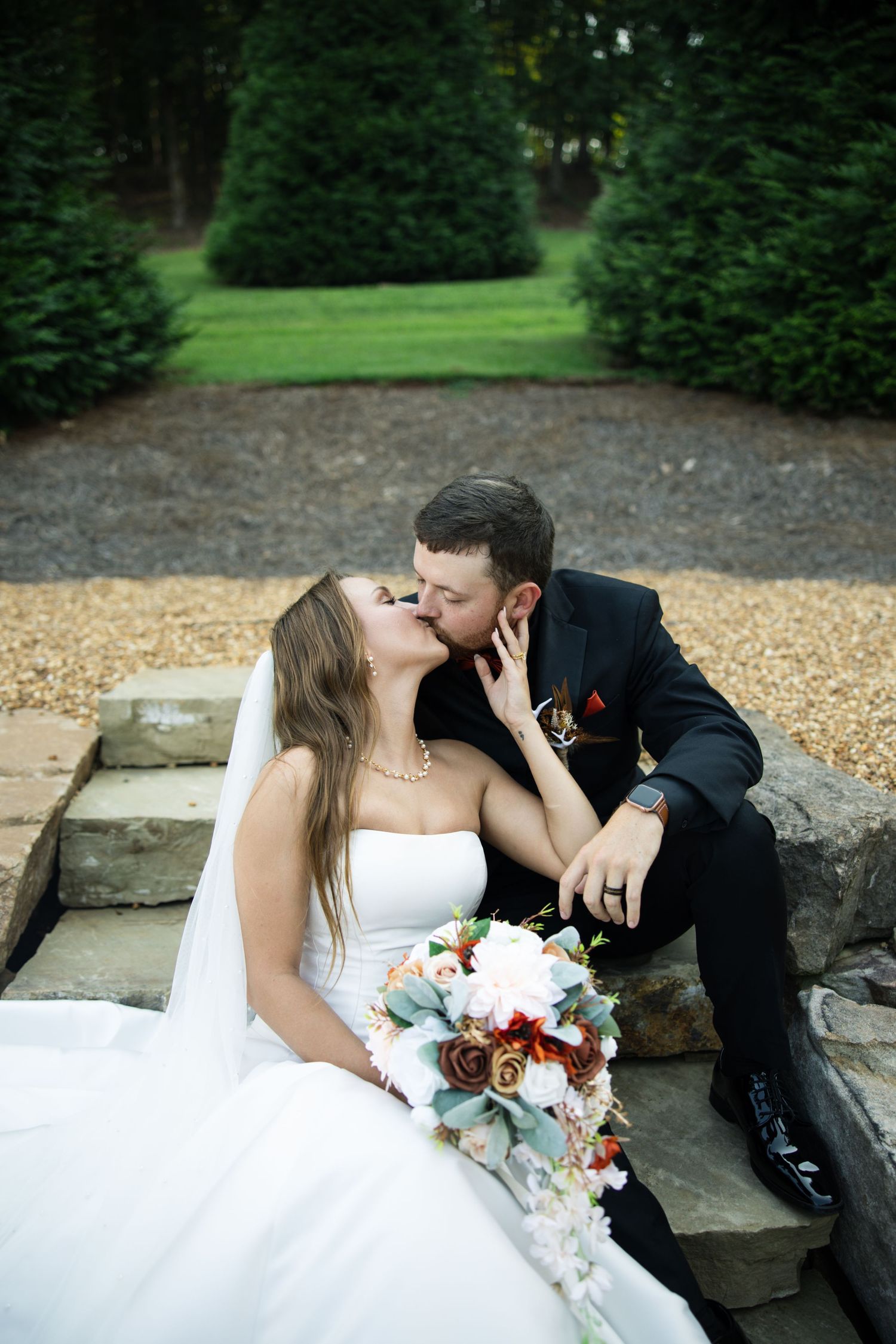 Bride and groom share a romantic kiss on stone steps surrounded by lush greenery, bride holding a beautiful floral bouquet.