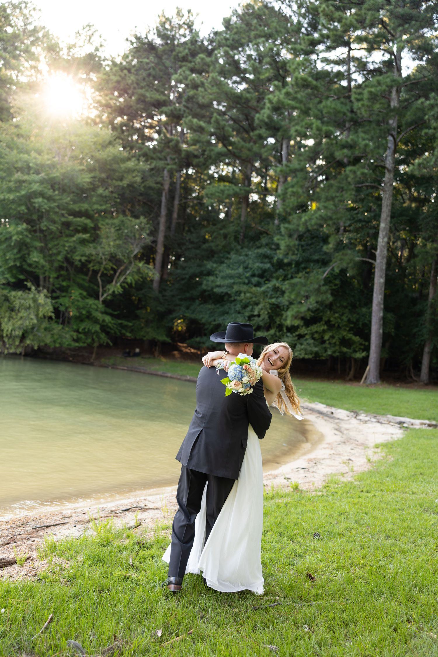 Bride and groom embracing near a serene lakeside with lush green trees and golden sunlight in the background.
