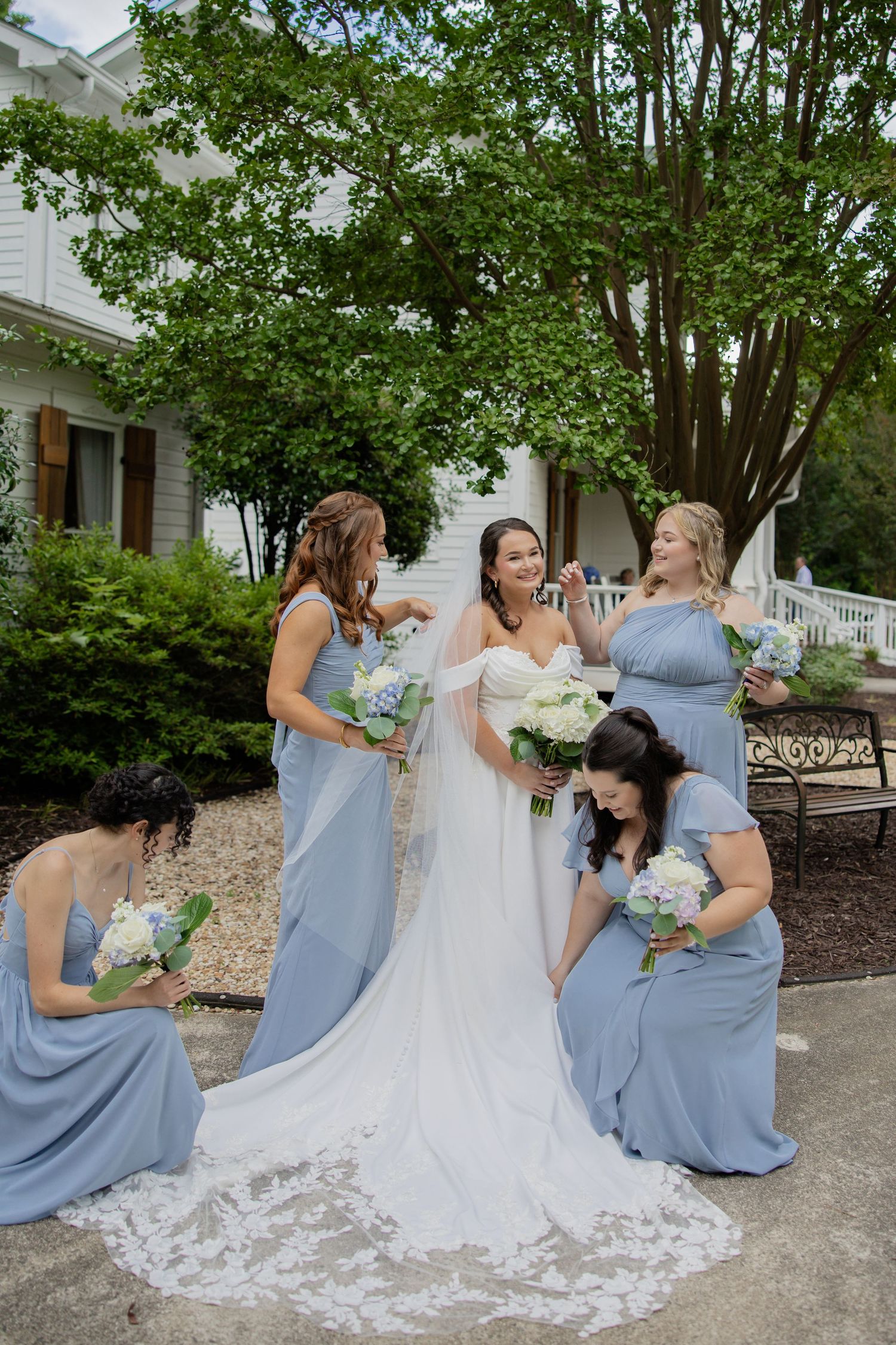 Bridesmaids in light blue dresses gather around a bride with a flowing white train outside a white house.