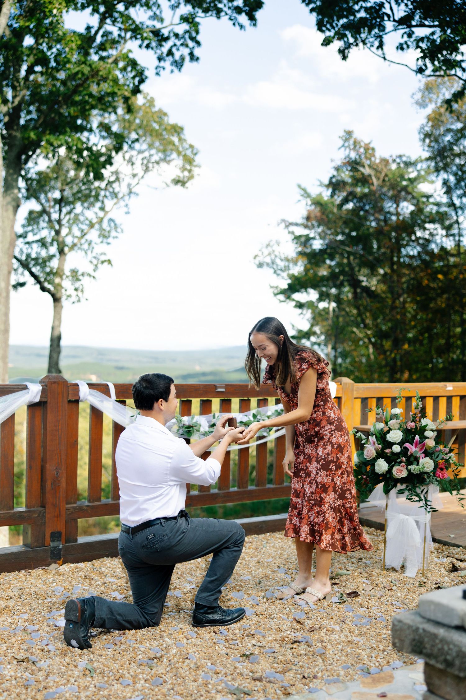 A romantic proposal moment on a scenic wooden deck overlooking mountains with flowers in a vase nearby.