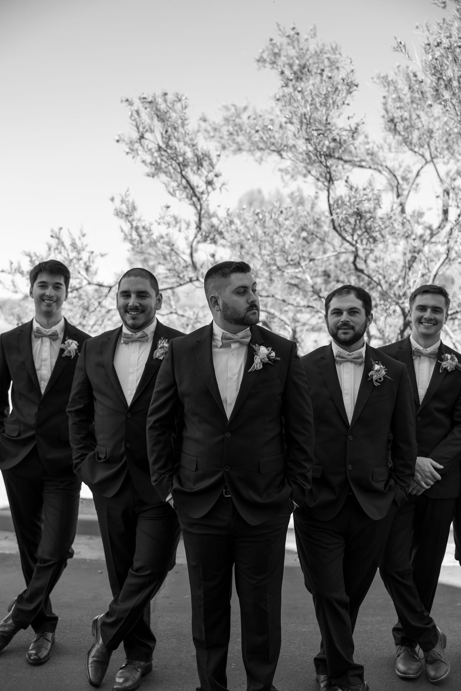 Black and white photo of groom and groomsmen in dark suits with bow ties and boutonnieres posing outdoors.