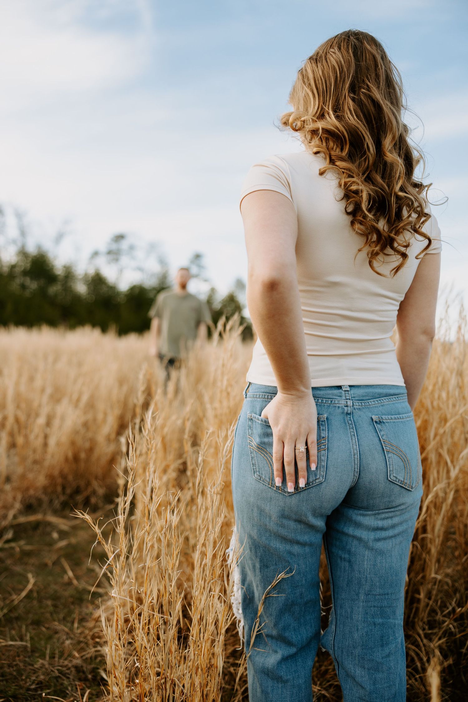 A woman in jeans looks towards someone in the distance across a golden field.
