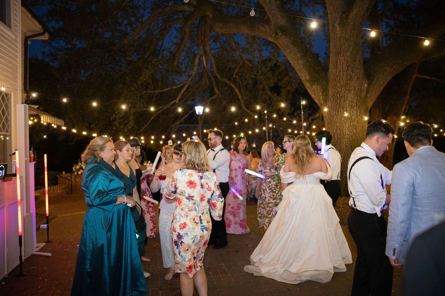 Wedding guests dance under twinkling string lights at an outdoor evening reception with trees overhead.
