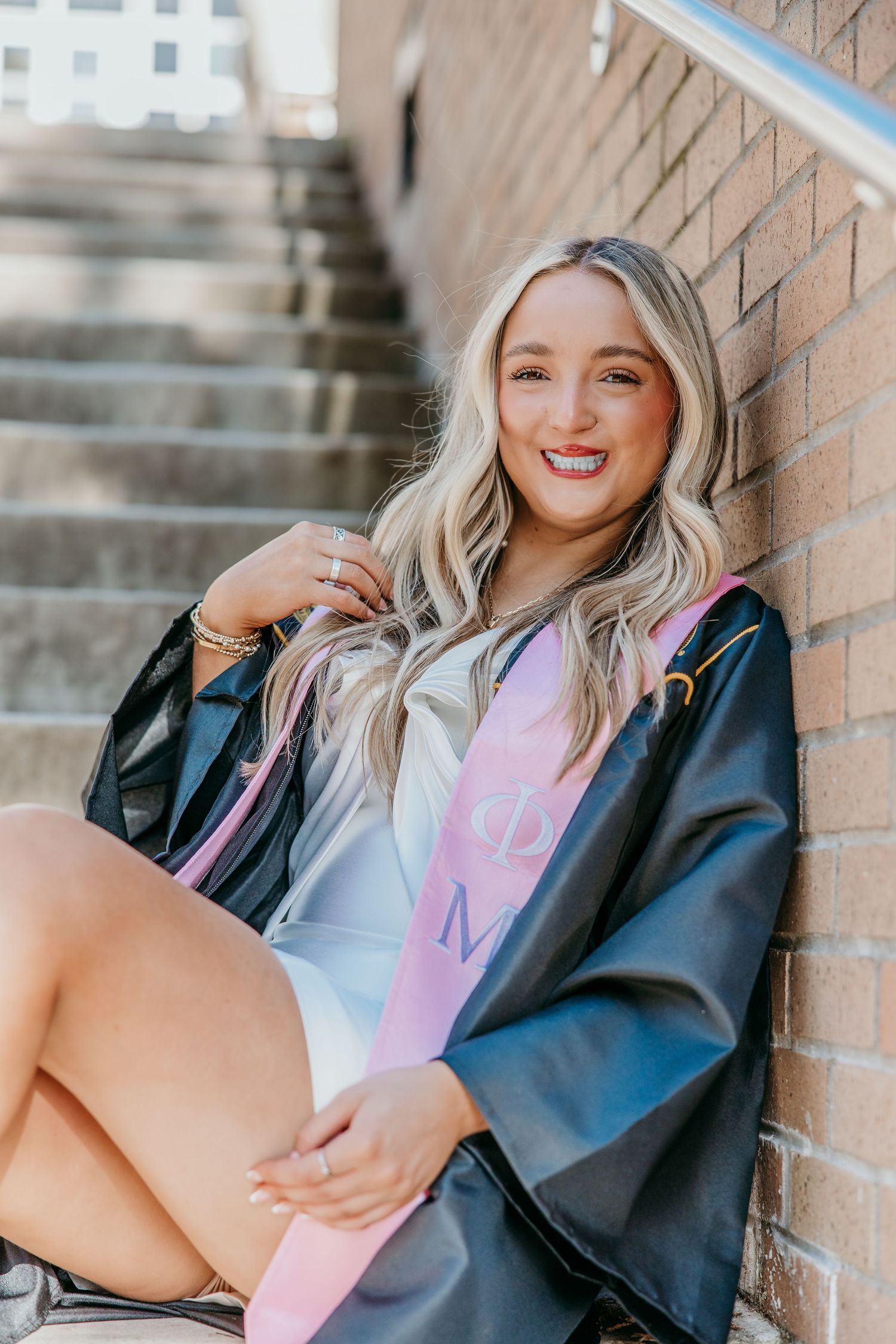 Graduate in cap and gown sitting against brick wall on stairs.
