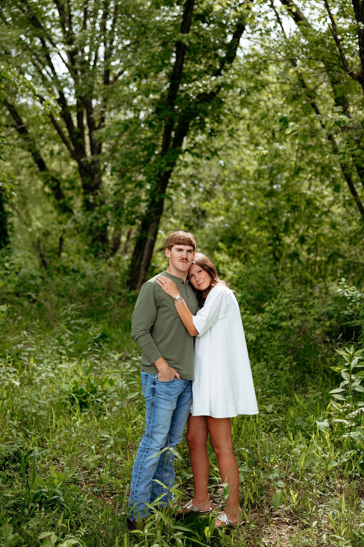 A couple embraces in a lush green forest, woman in white dress leans on man in green shirt and jeans.