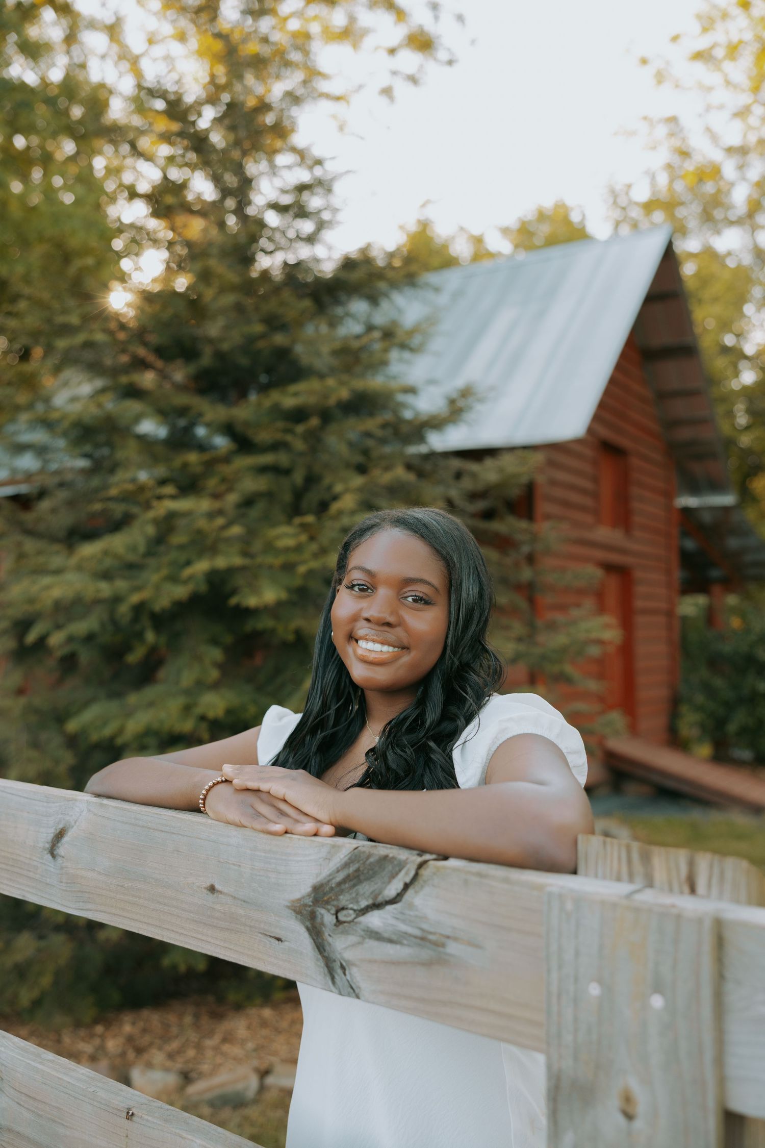 Someone in white leans on a wooden fence with a rustic cabin in the background.