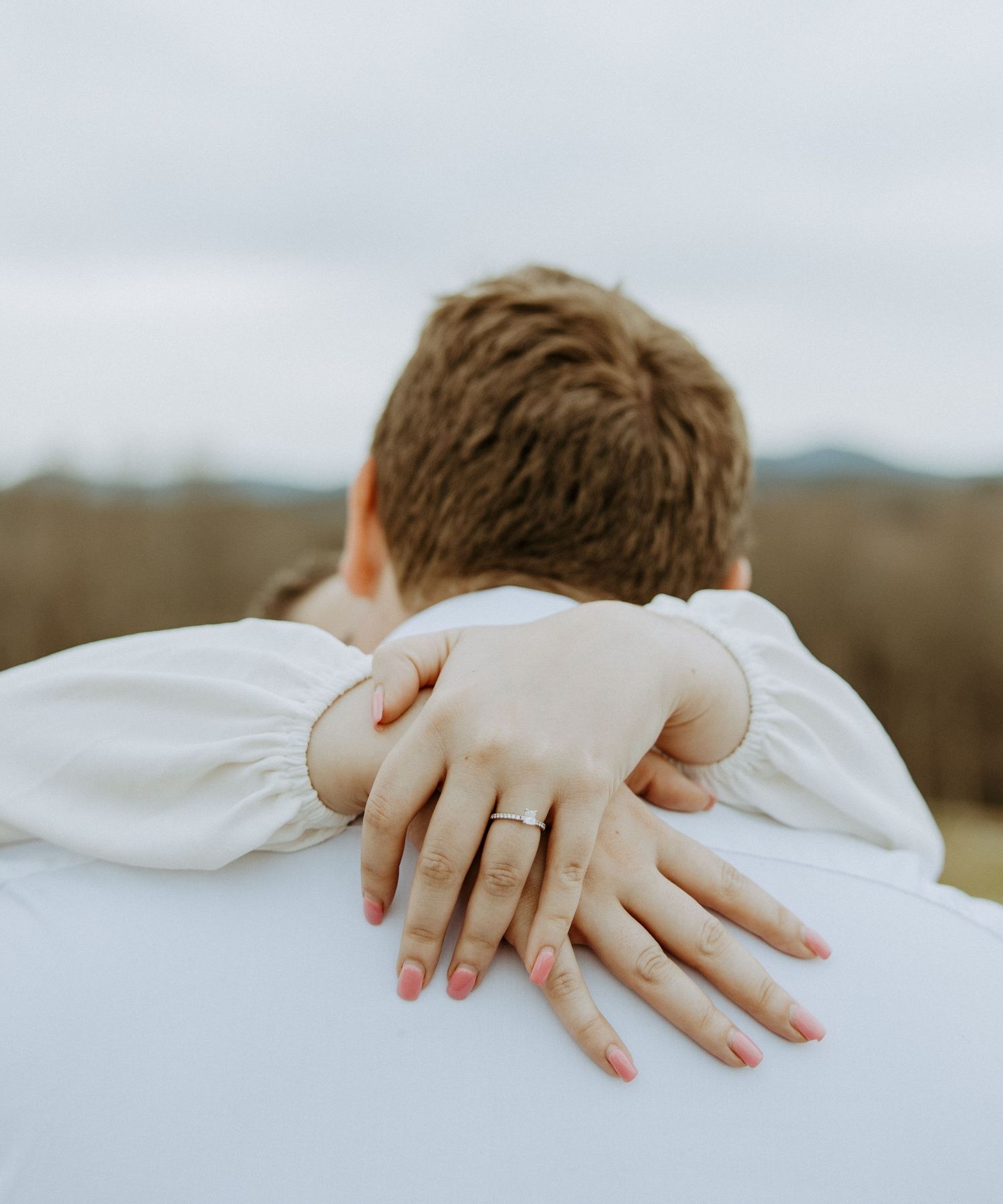 Close up of hands embracing from behind with an engagement ring visible against white clothing.