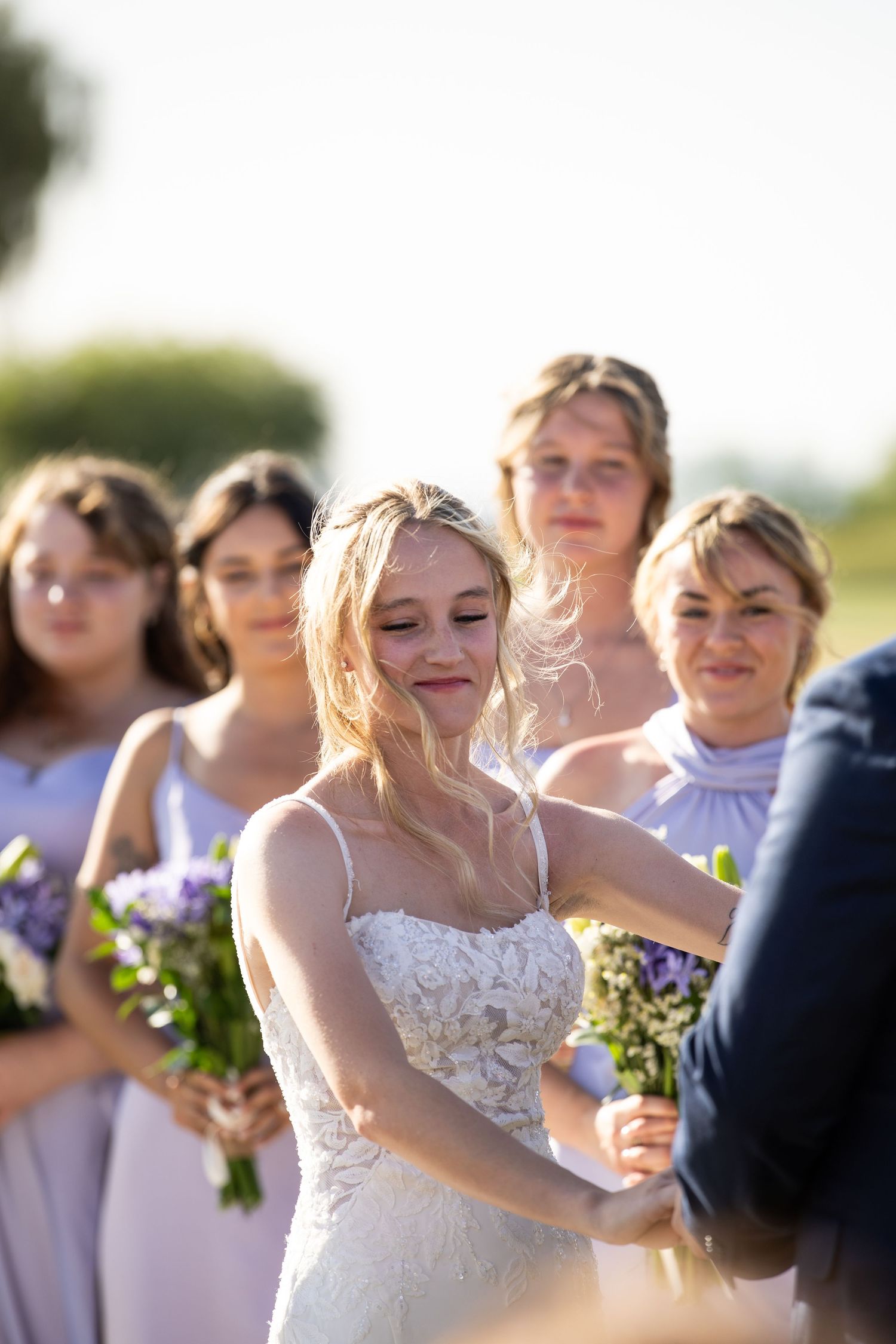 Smiling blonde bride in lace gown holding bouquet during outdoor wedding ceremony.