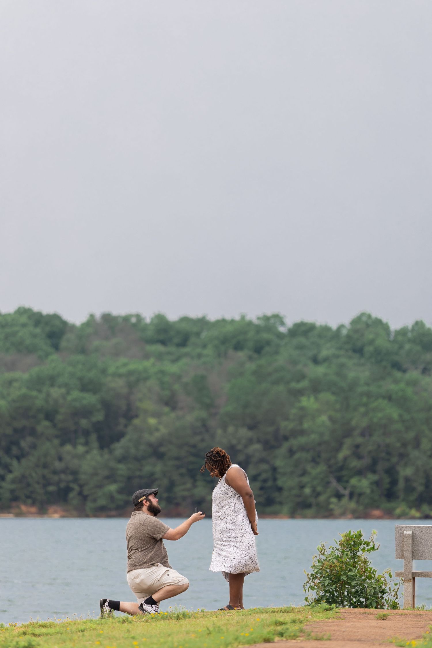 Two people sit on a grassy hill overlooking a peaceful lake surrounded by trees on a cloudy day.